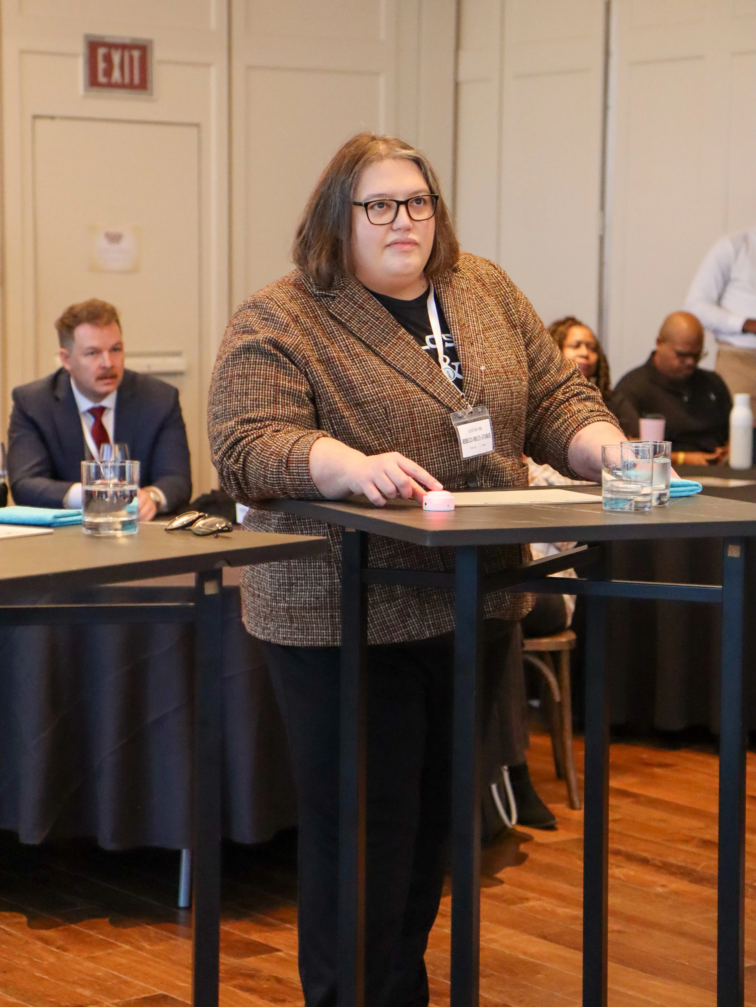 A woman with shoulder-length brown hair and glasses stands behind a table in a conference room, wearing a brown checkered blazer. Other people are seated and standing in the background.