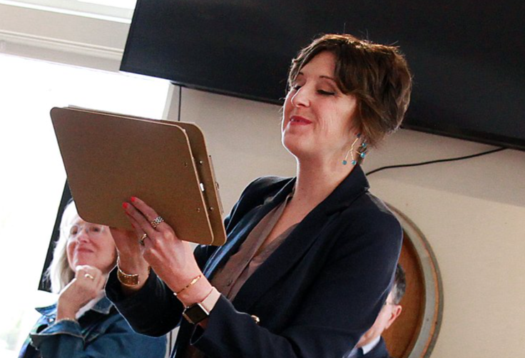 A woman in a black blazer is holding and looking at a brown clipboard, smiling with her eyes closed, in a room with a large flat-screen TV and a wooden-framed mirror in the background.