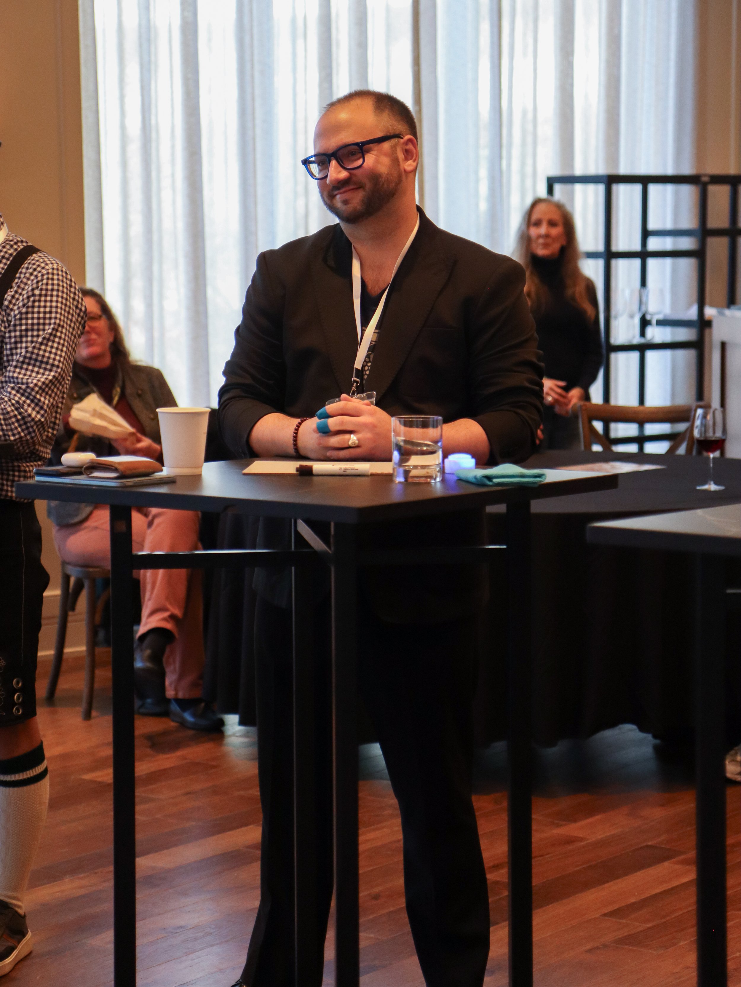 A man with glasses and a beard sitting at a table during a meeting or conference, with other people in the background.