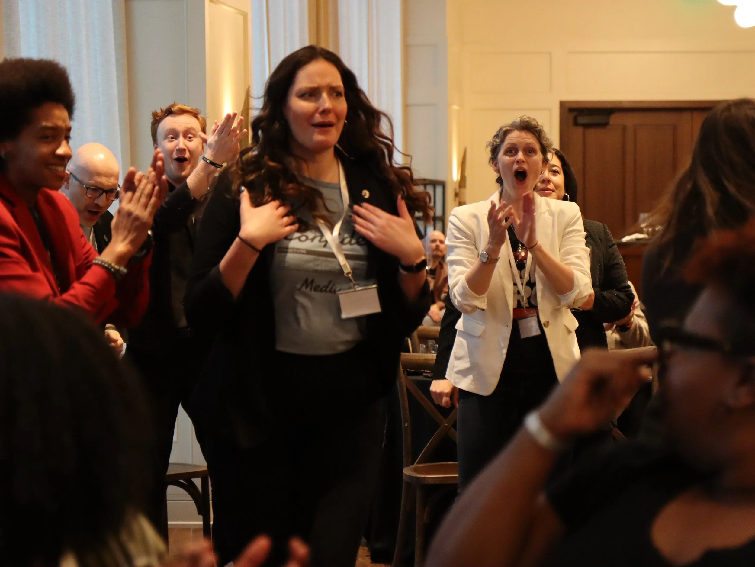 A group of diverse people at an indoor event, some with expressions of surprise or emotion, one woman with her hand on her chest, others clapping or reacting enthusiastically.