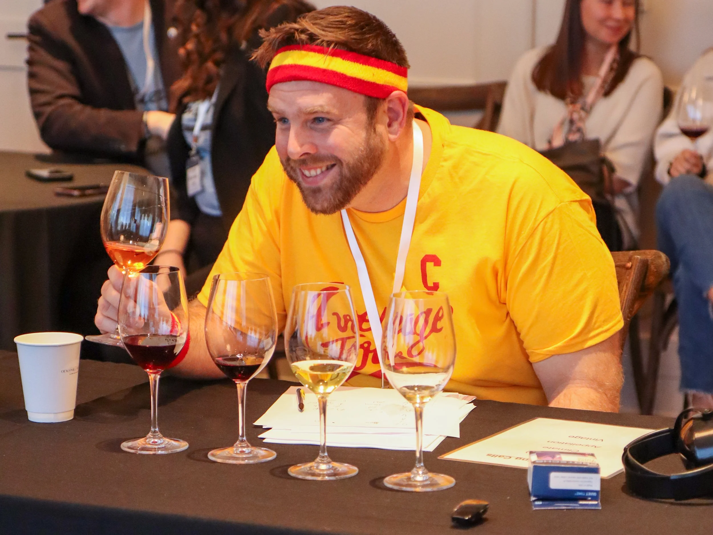 A man with a beard, wearing a yellow shirt and a red and yellow headband, is smiling and holding a glass of rosé wine at a table with multiple wine glasses, papers, and a cellphone. In the background, two women are sitting and smiling, one with glass