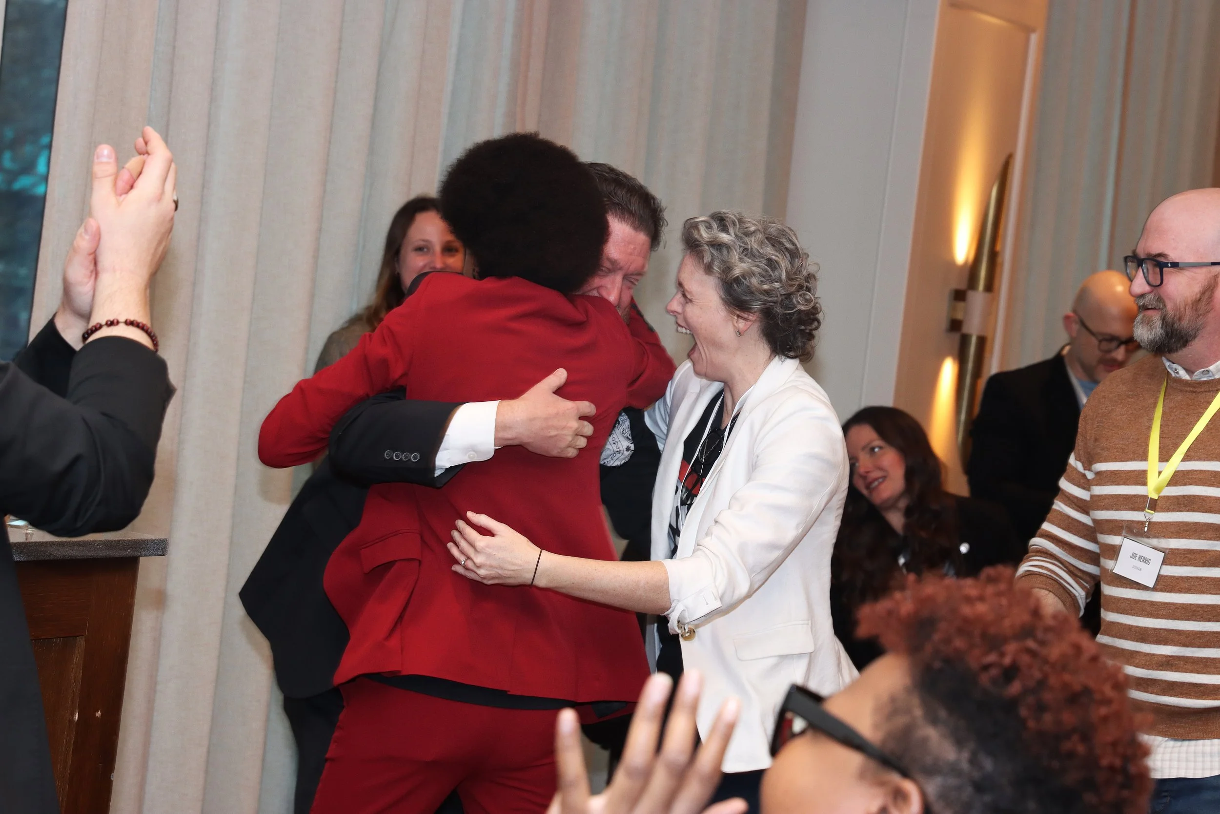 People at an event hugging and smiling, with some looking on and others taking pictures.
