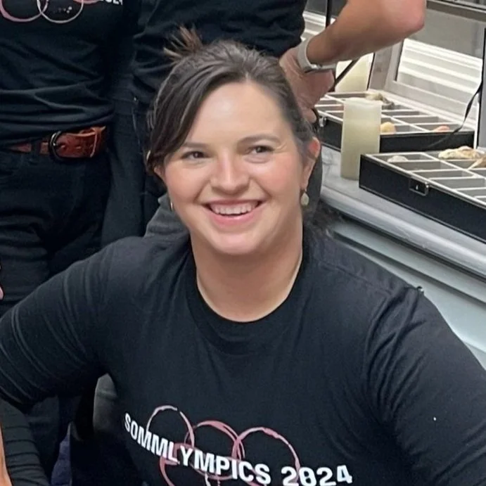 Smiling woman wearing a black "SOMMOMYLPICS 2024" T-shirt, seated at a table with jewelry display trays in the background.