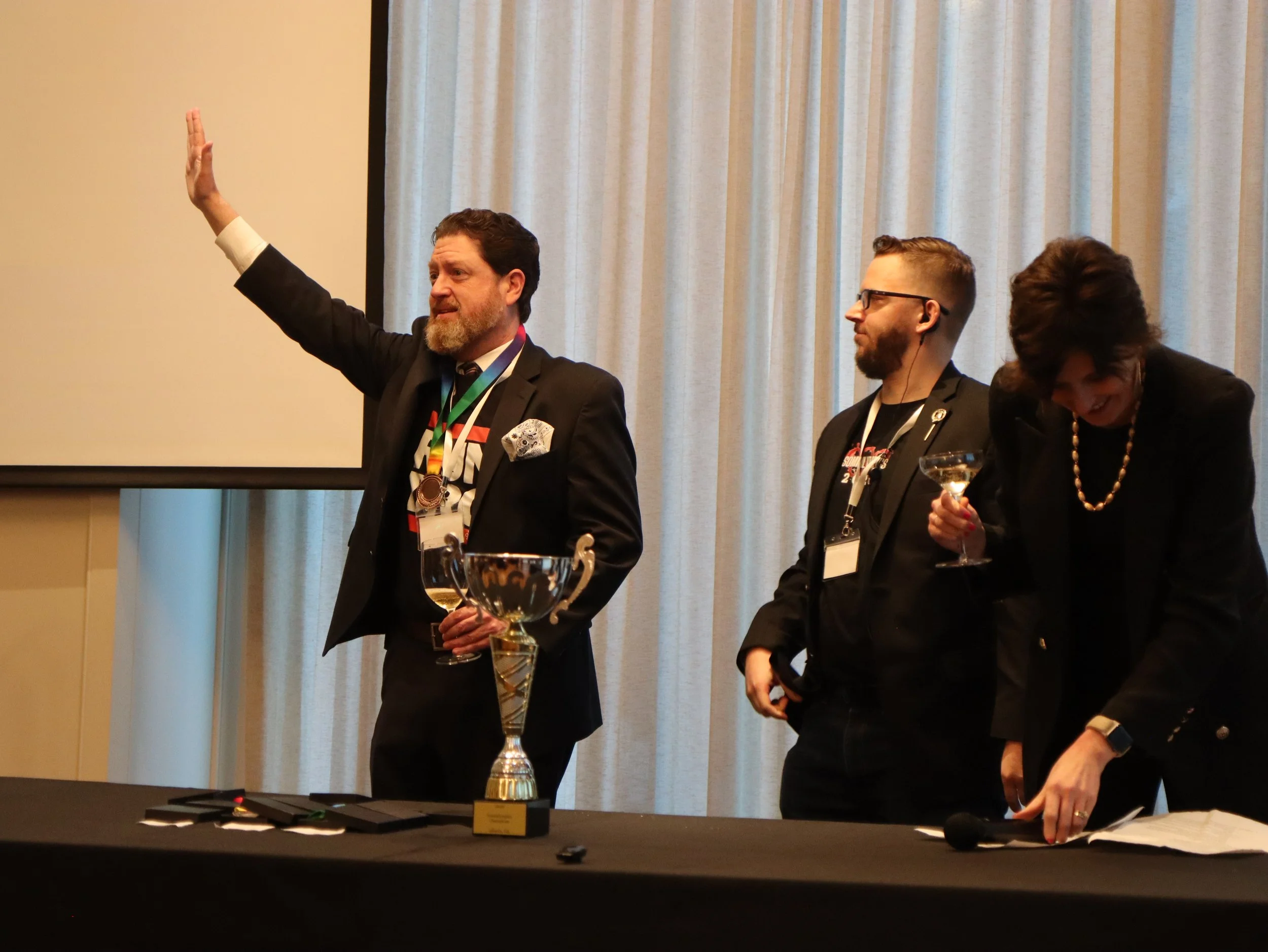 A man with a beard and glasses raises his hand during a formal event, standing next to a woman with frizzy hair and a pearl necklace, both wearing dark suits. They are at a table with trophies and papers, with a woman smiling and holding a drink near