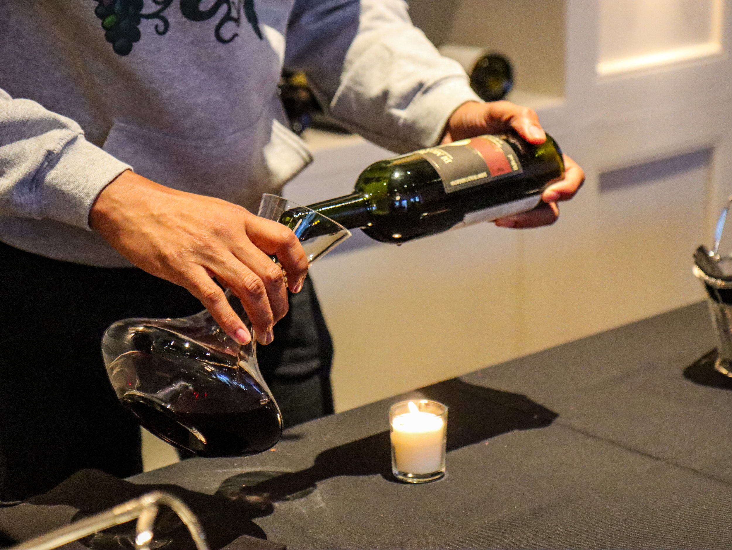 Person pouring red wine into a glass over a table with a candle.