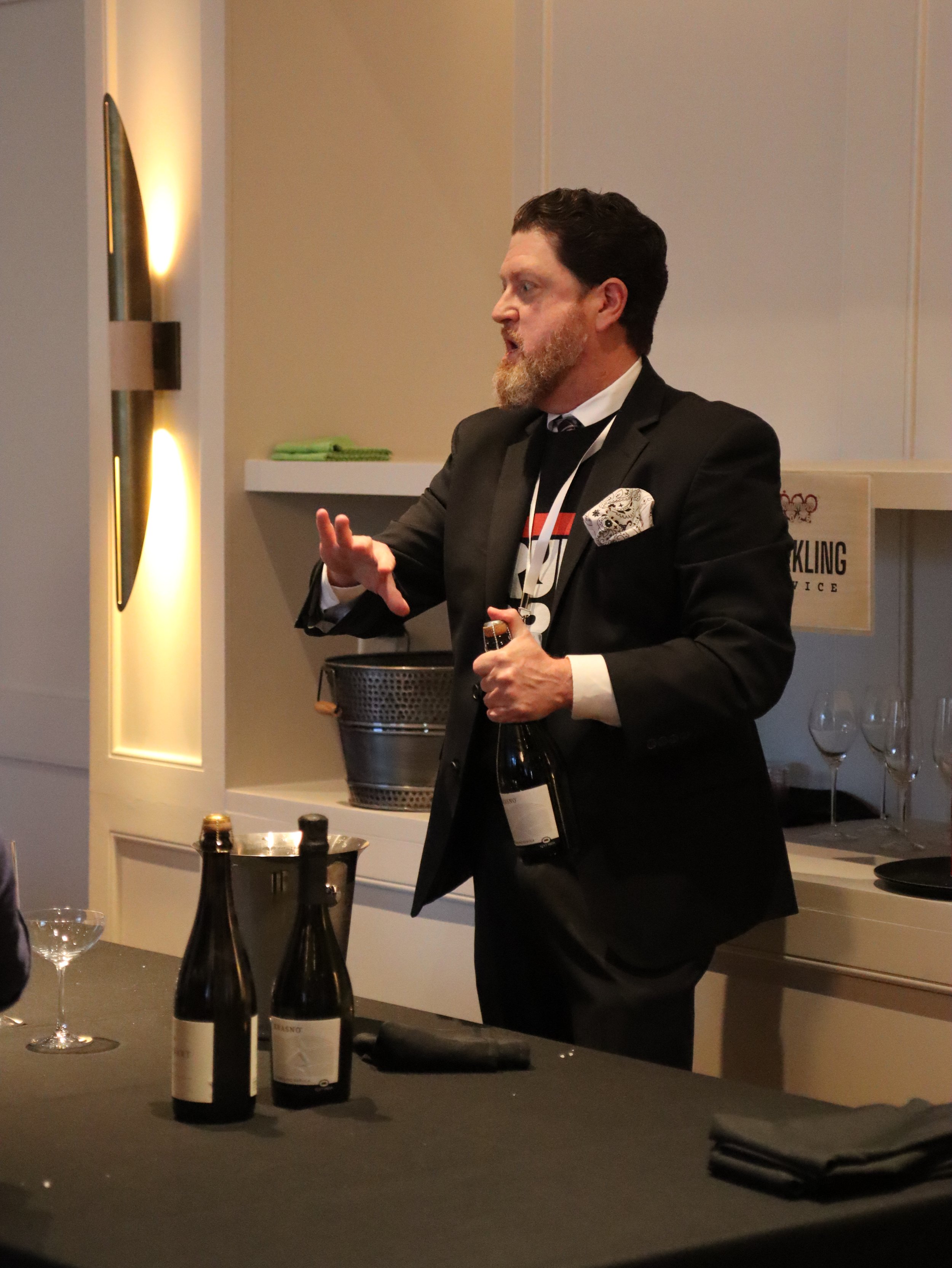 A man in formal attire gestures with his hand while holding a bottle of wine, standing in a room with wine bottles, glasses, a basket, and a sign that says "Book Club" in the background.