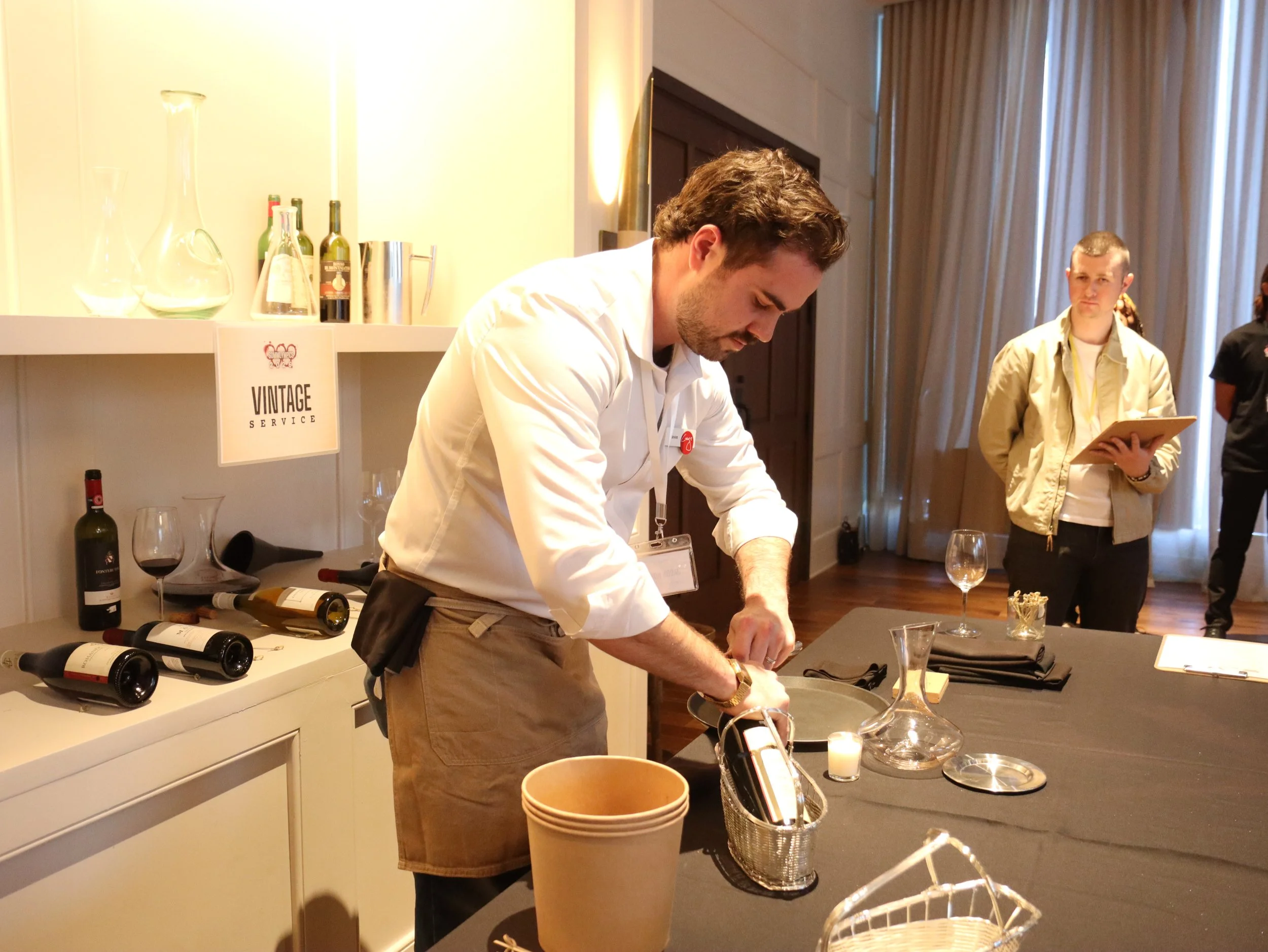Man pouring wine at a tasting event with wine bottles and glassware on the table, and two women in the background, one holding a notebook.