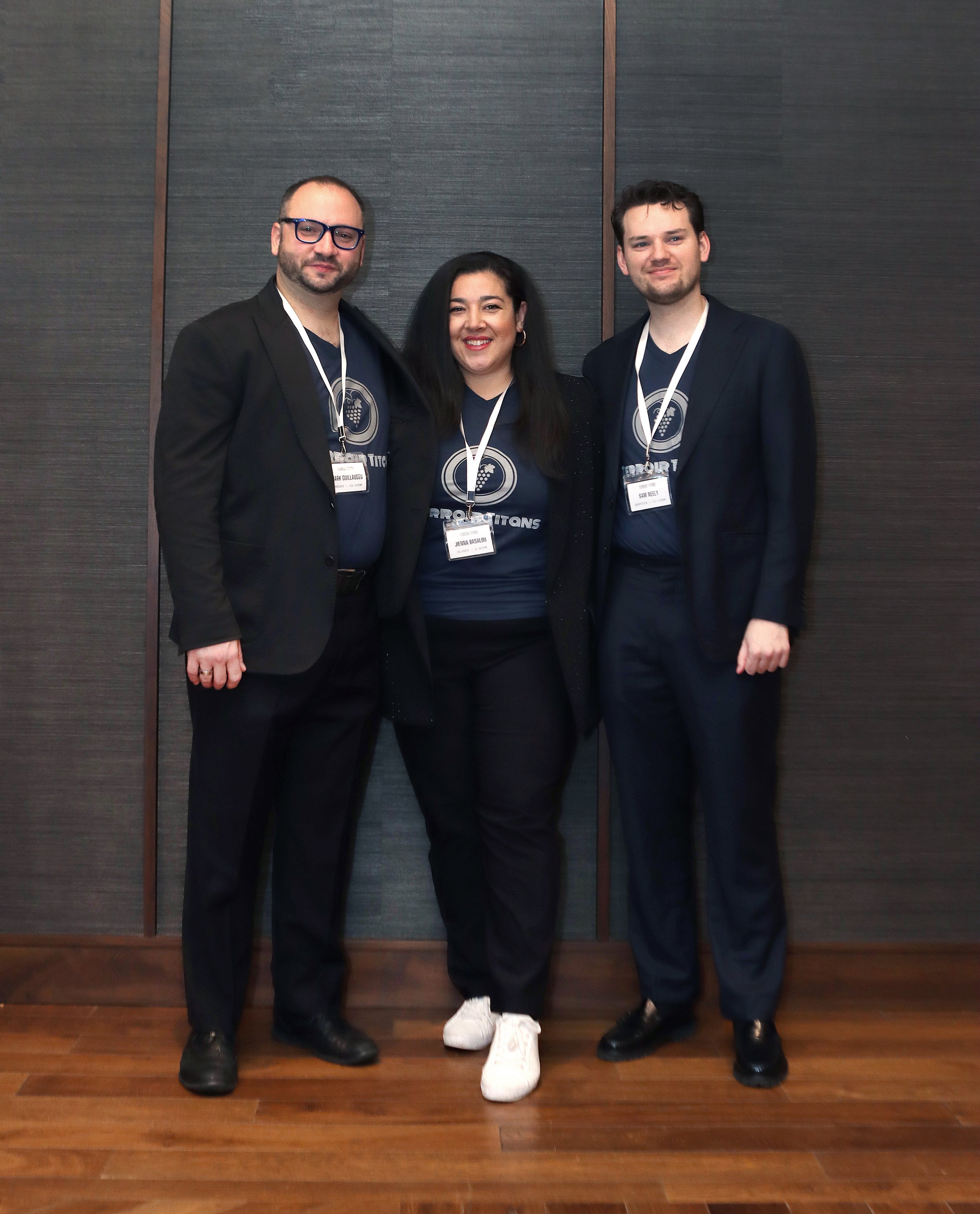 Three people, two men and one woman, standing together indoors against a dark wall, all wearing dark blazers and blue shirts with event badges around their necks. The woman is wearing white shoes, while the men are in black shoes.