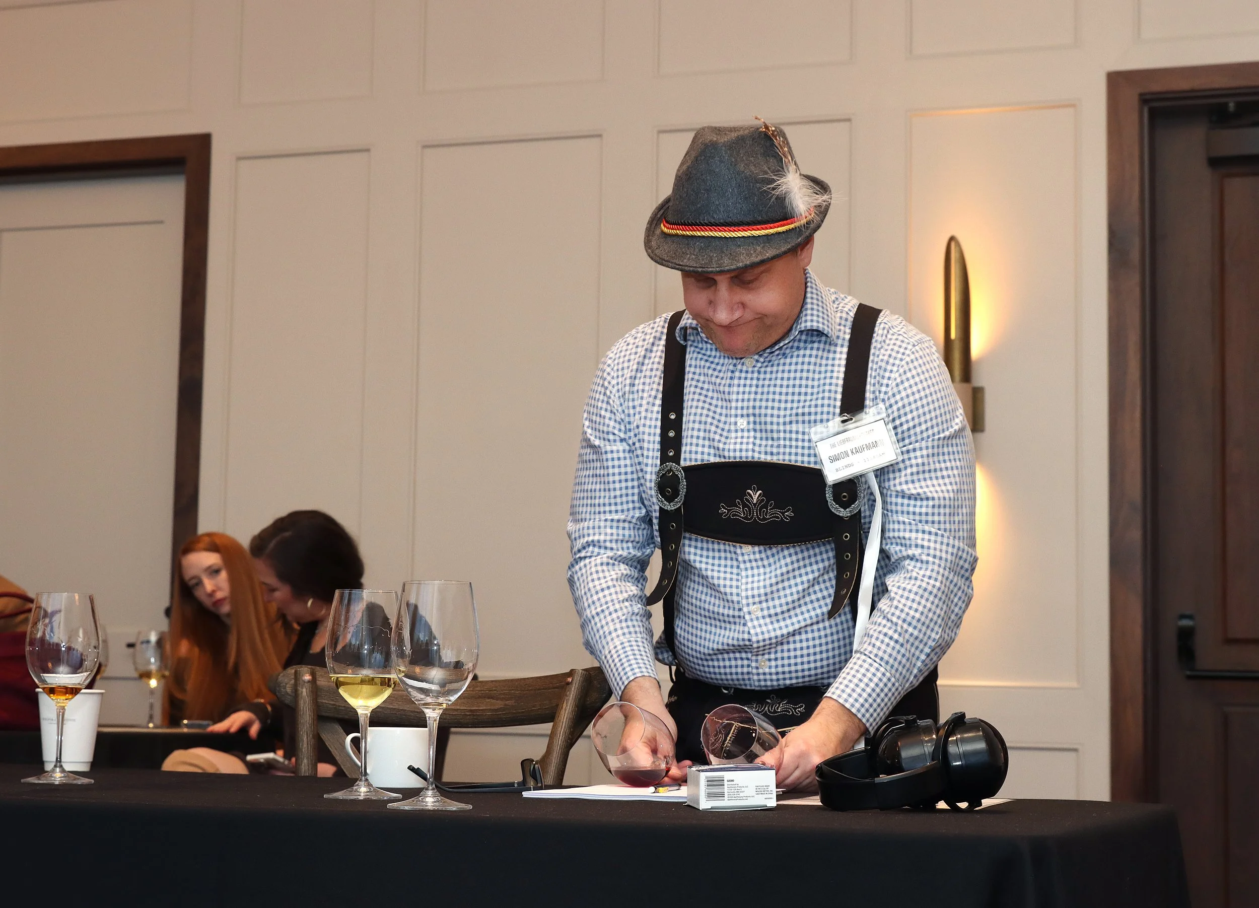 A man wearing a traditional Bavarian hat with a feather, checkered shirt, and apron, pouring wine at a table in a formal setting. There are wine glasses on the table, and several women are seated nearby, with some looking at their phones. The backgro