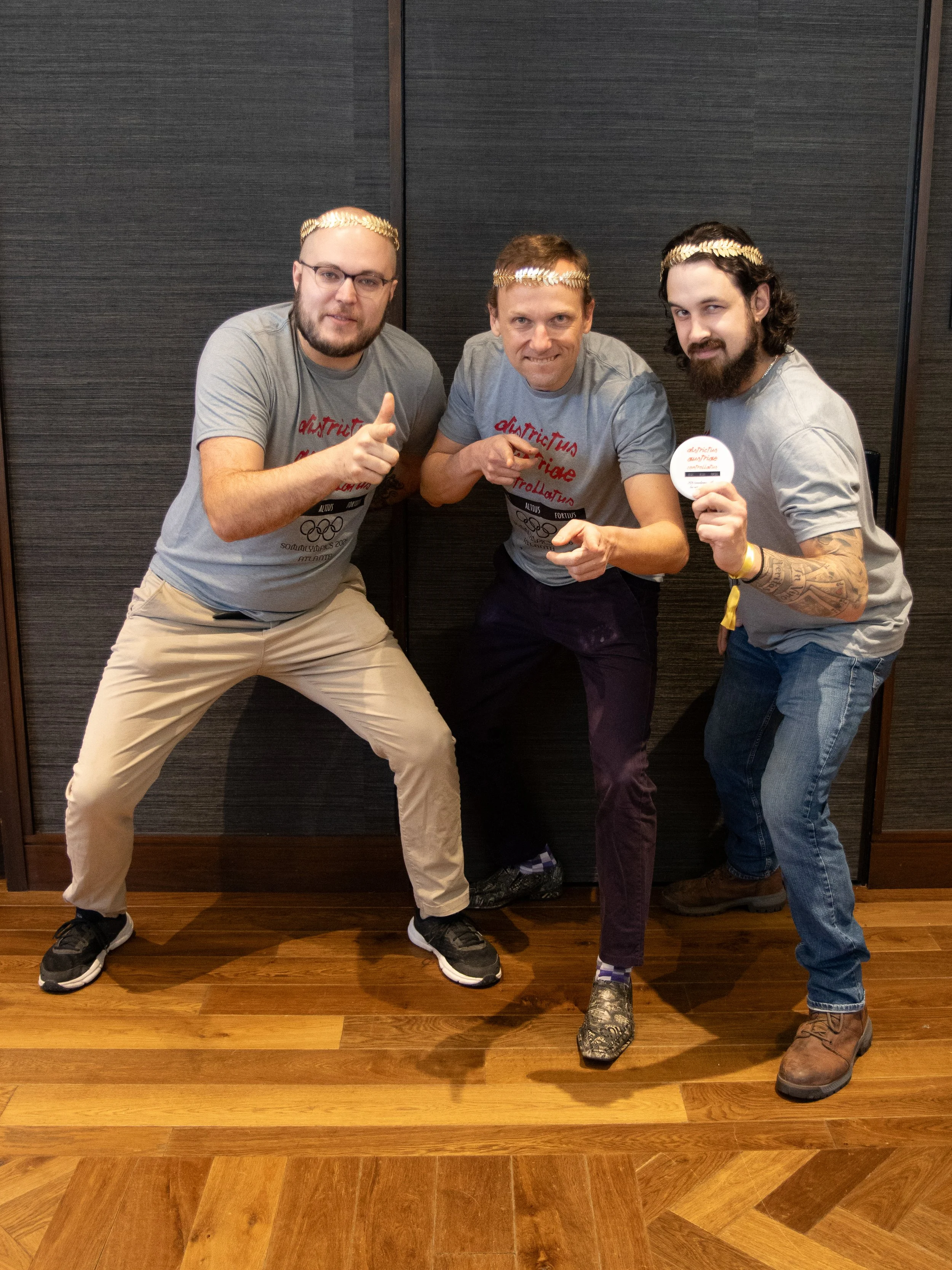 Three men wearing gray shirts and laurel leaf headbands posing with energetic gestures in front of a dark wooden wall.