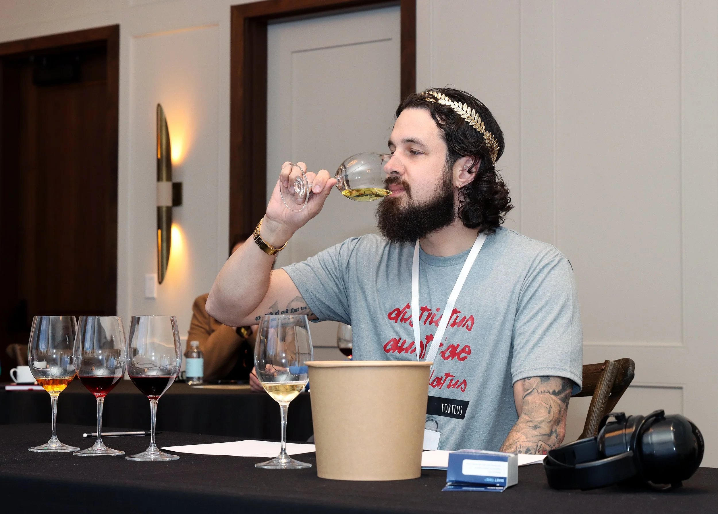 A man with long dark hair, a beard, and tattoos on his arms is tasting wine at a table with several glasses of wine. He is wearing a light gray T-shirt with red text, a gold leaf headband, and a gold watch. On the table are multiple wine glasses fill