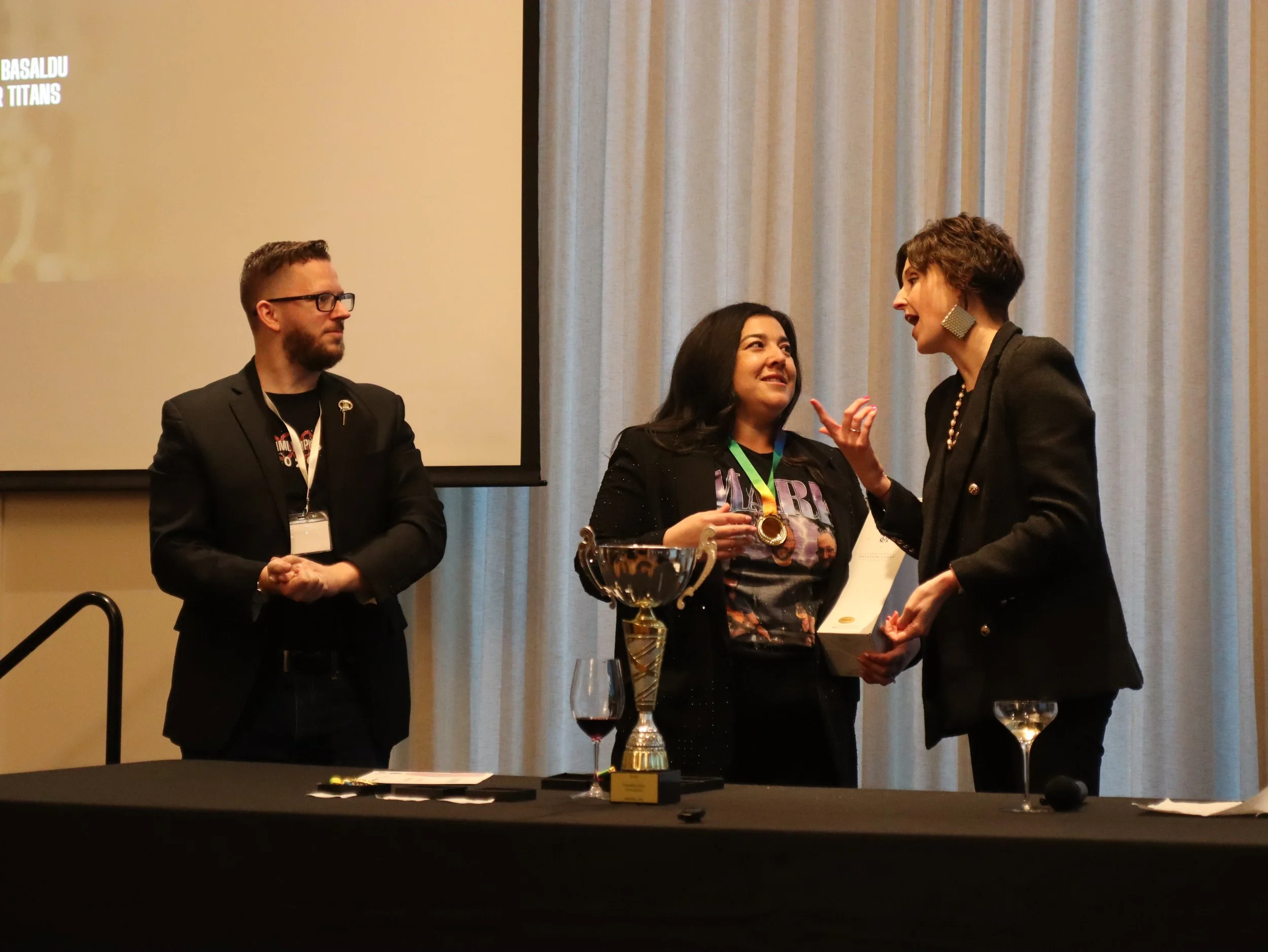 Three women and one man at an award ceremony, with a large screen listing names behind them. The women are smiling, and one is wearing large earrings and speaking to another woman. A table with a trophy and glasses is in the foreground.