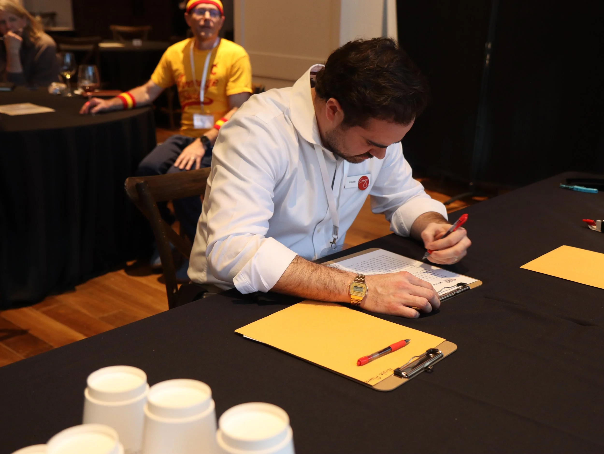 A man with dark hair and a beard writing on a yellow piece of paper at a table, with a man in a yellow shirt and glasses sitting behind him, and a woman sitting at a different table in the background. There are pens, a clipboard, and bottles in the f