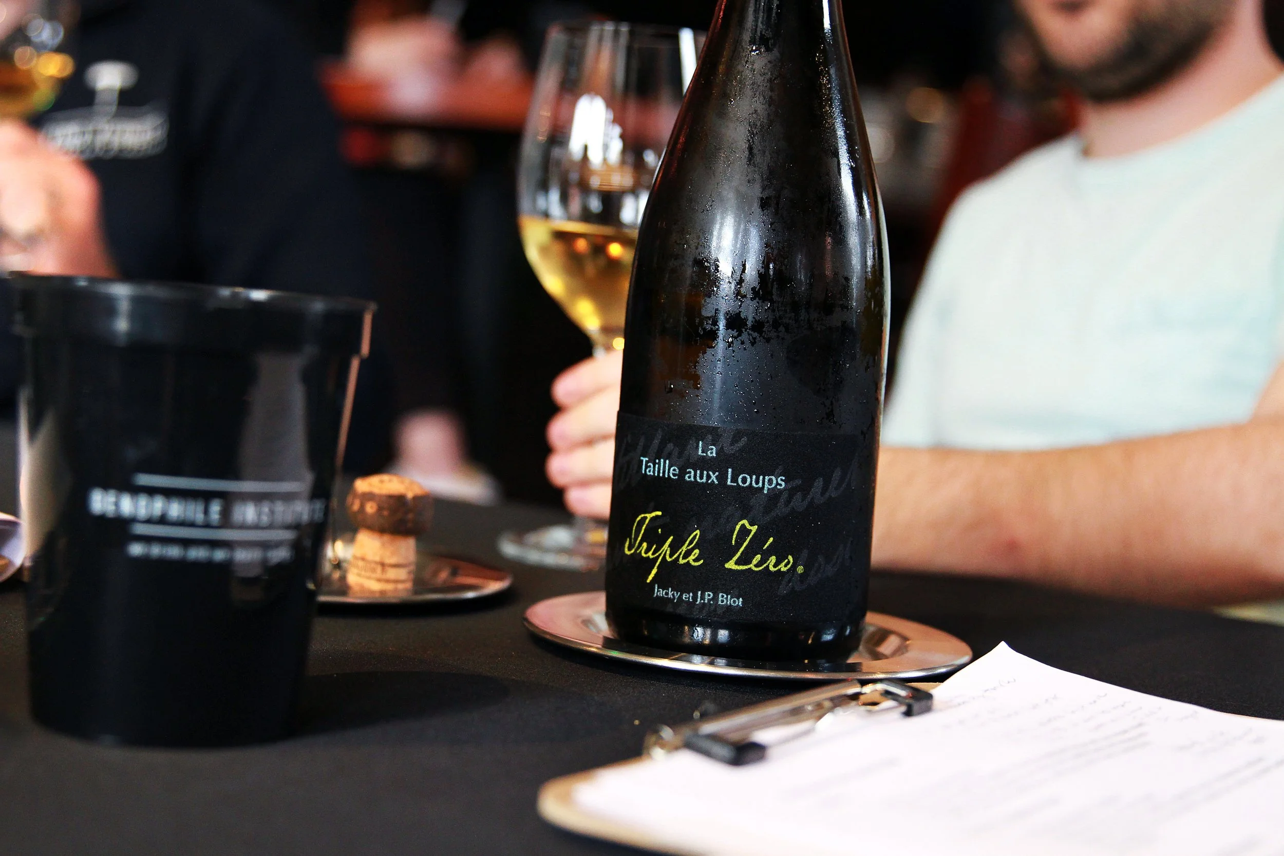A bottle of wine labeled 'Triple Zero' and 'Taille aux Loups' on a table along with a glass of white wine, a black pen, and some small desserts in a wine tasting setting.
