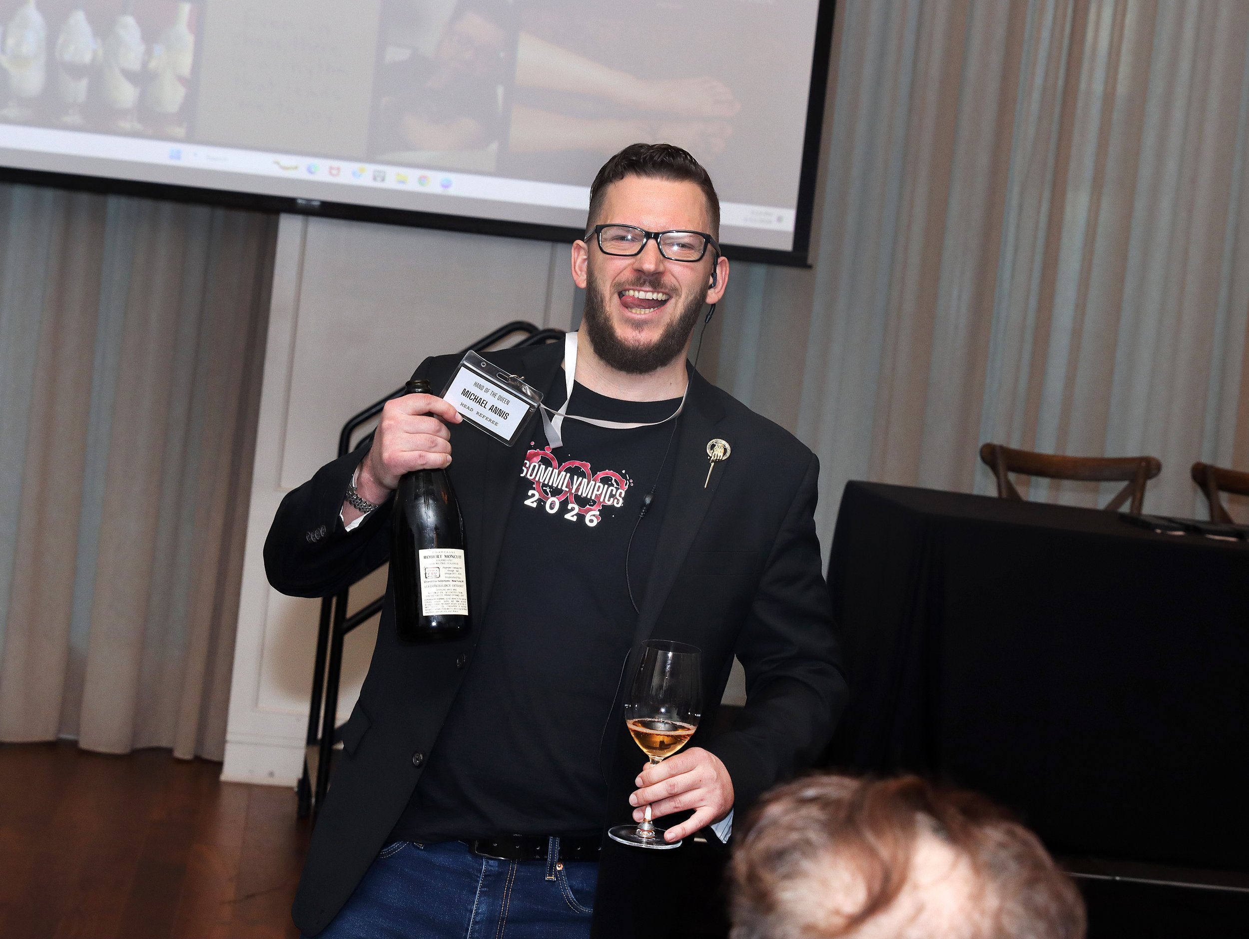 A man with glasses and a beard wearing a black jacket and a 'Sommy Olympics 2026' t-shirt, smiling and holding a wine glass with a pink drink, standing in a room with a large television screen behind him.