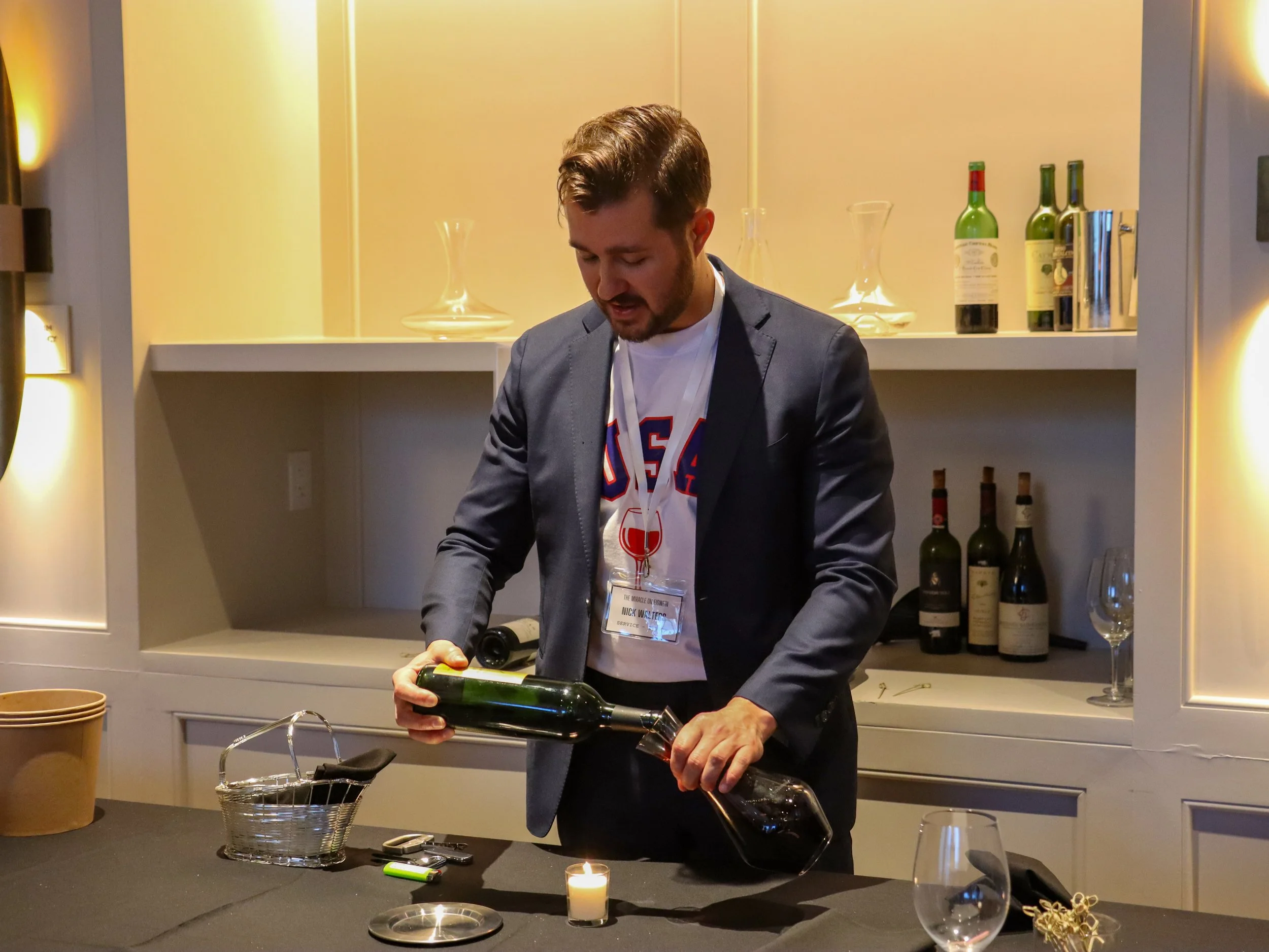 A man pouring red wine from a bottle into a glass at a table with a small lit candle, wine bottles, and glassware in the background.