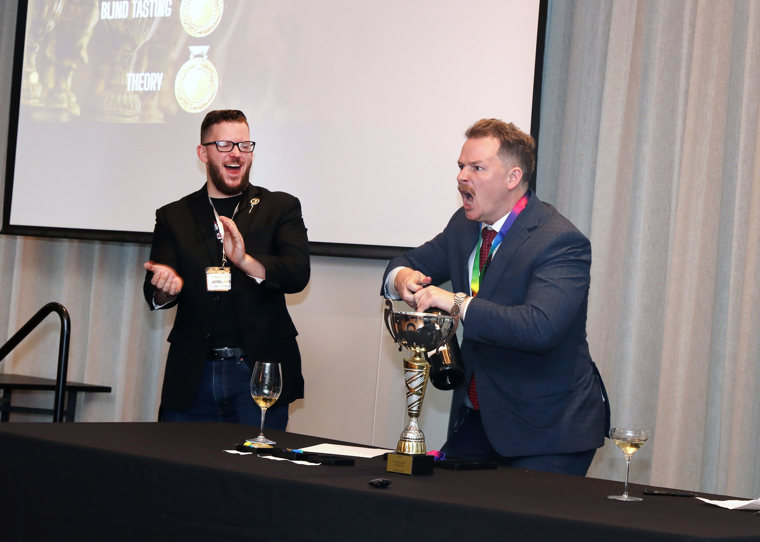 Two men in suits are at a table with trophies, glasses, and papers, celebrating during an award ceremony. One man is smiling and clapping, the other appears excited and is speaking or cheering.