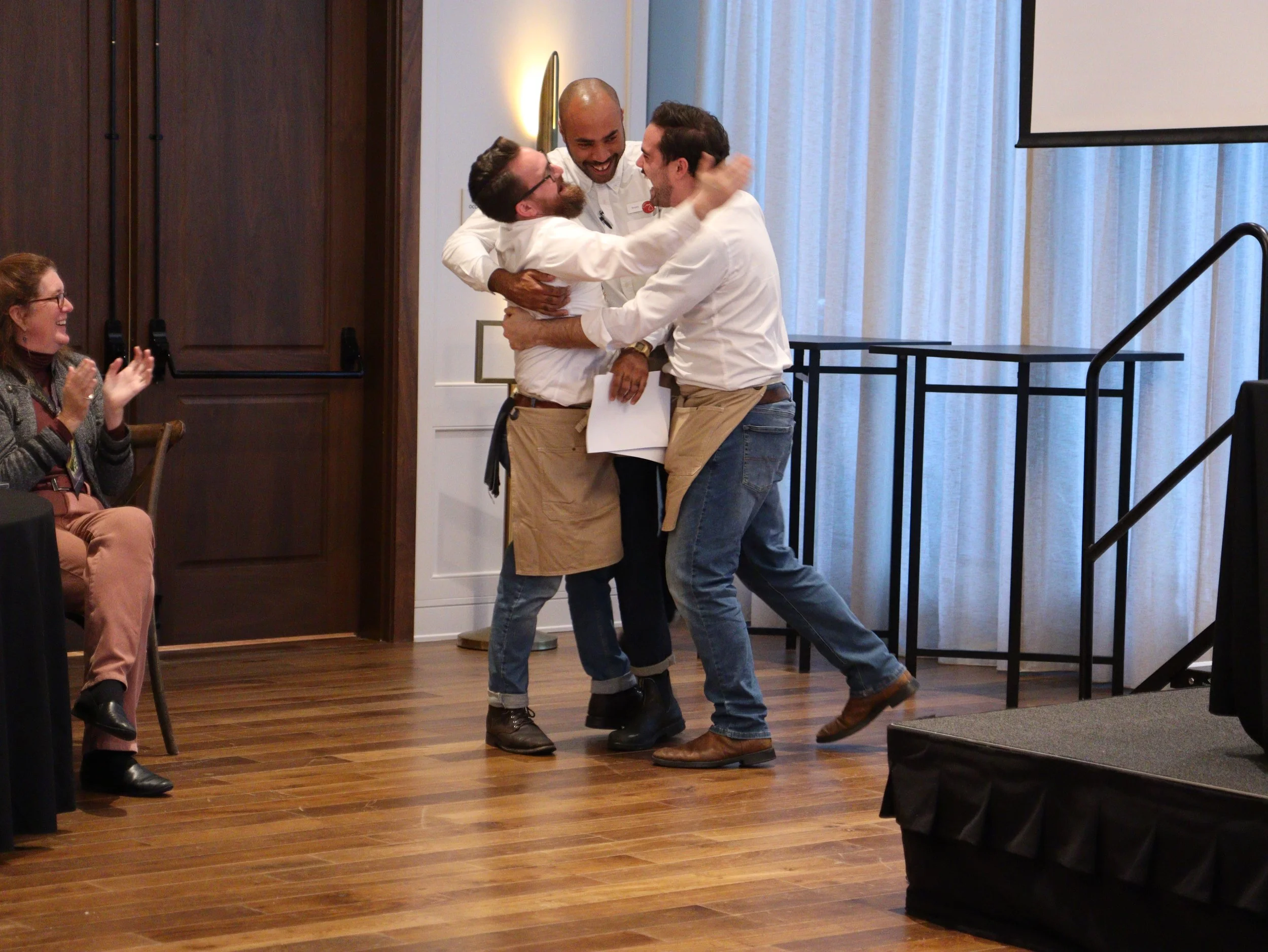 Three men are joyfully celebrating in the center of a room, hugging and smiling. A woman on the left is clapping and smiling. The room has wood flooring, a staircase railing, and curtains covering a window or wall behind them.