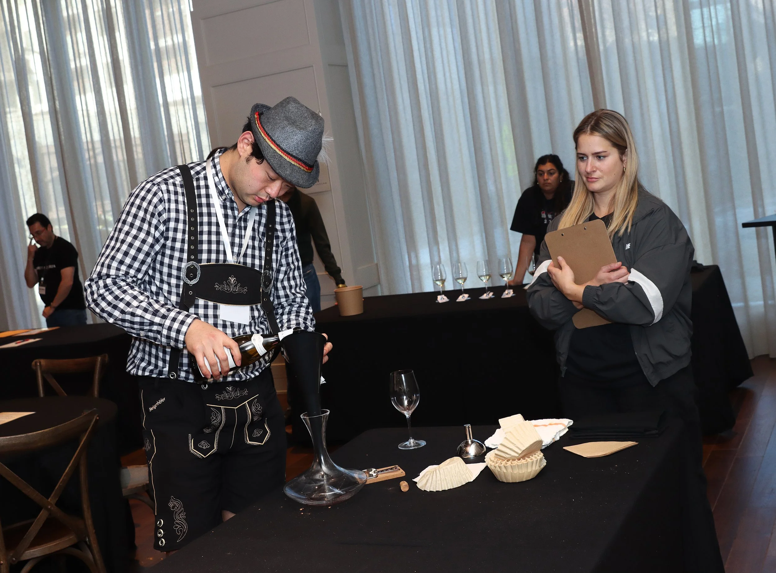 A person pouring wine into a glass at a tasting event, with others observing, set up with wine glasses and cheese on the table, and sheer curtains in the background.
