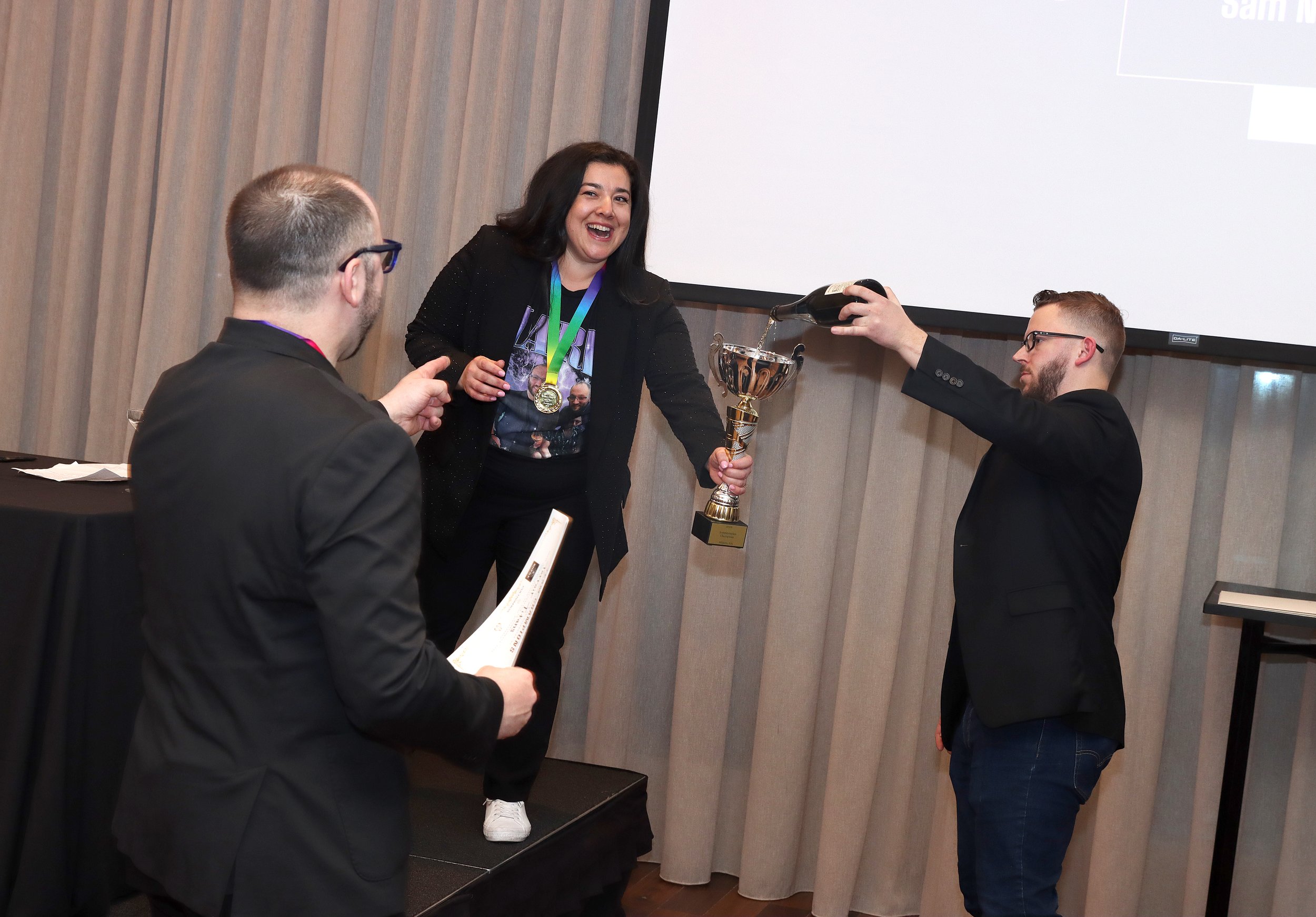 A woman with dark hair and a black jacket is standing on a podium, smiling and holding a trophy. She is wearing a medal around her neck. Two men are next to her, one holding a document and the other pouring champagne over her, celebrating her achieve