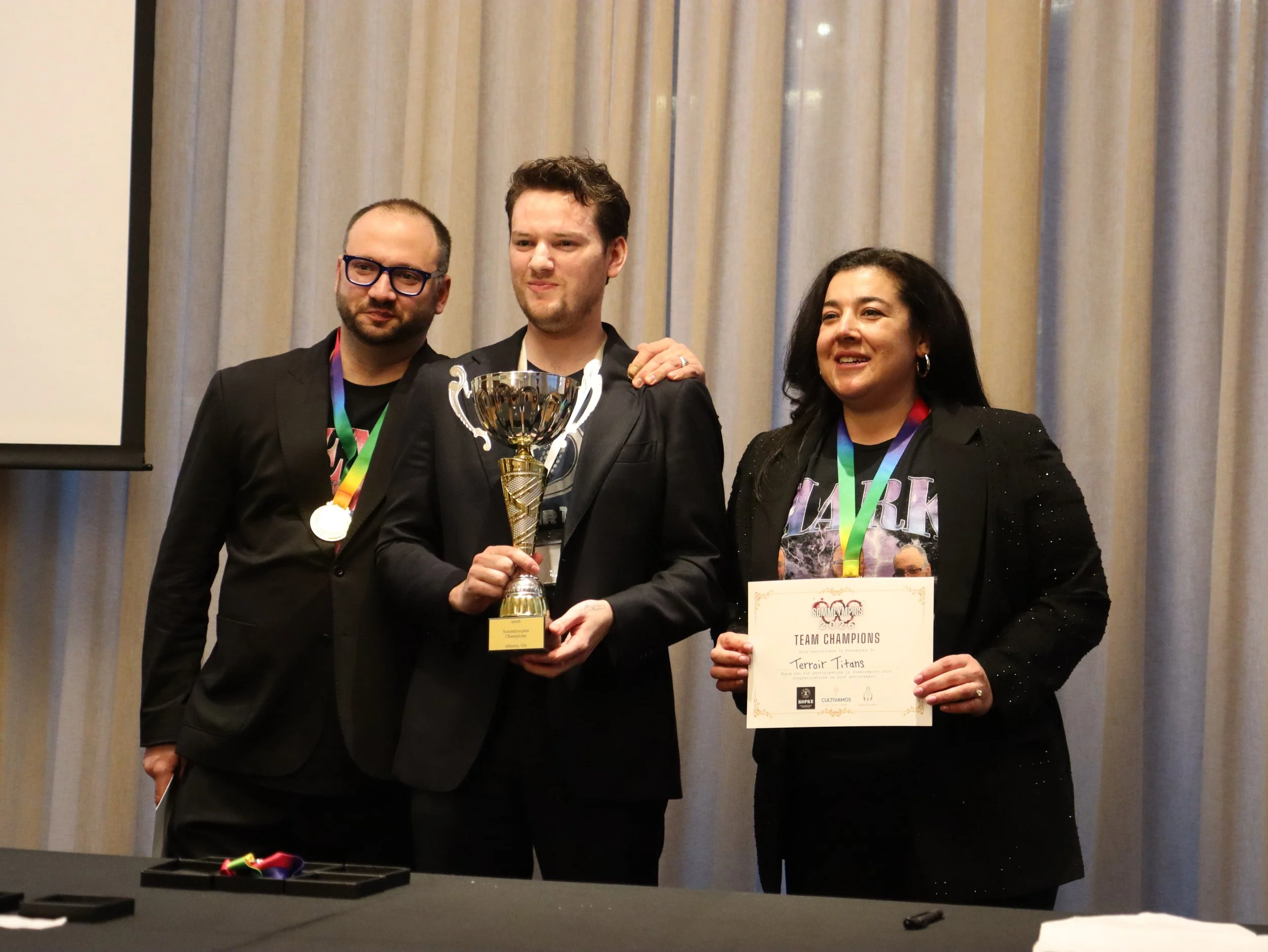 Group of four people at an award ceremony, two women and two men, smiling and holding awards and medals. They are in a room with framed maps and hanging lights.