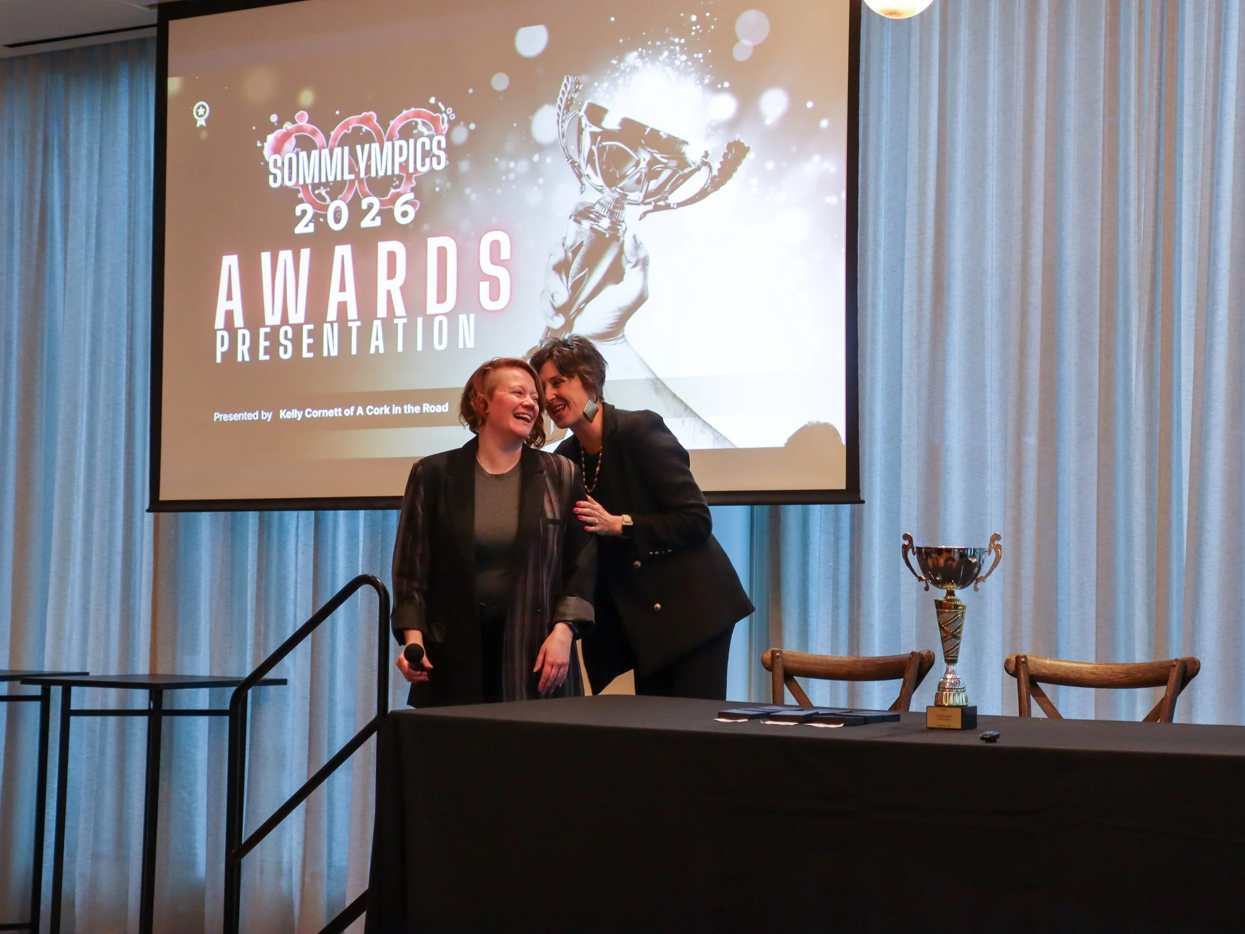 Two women standing together smiling in front of a stage with a large screen displaying an awards presentation. The screen shows a trophy graphic with the text 'Sommy Olympics 2026 Awards Presentation'. There is a table in front with a trophy and some