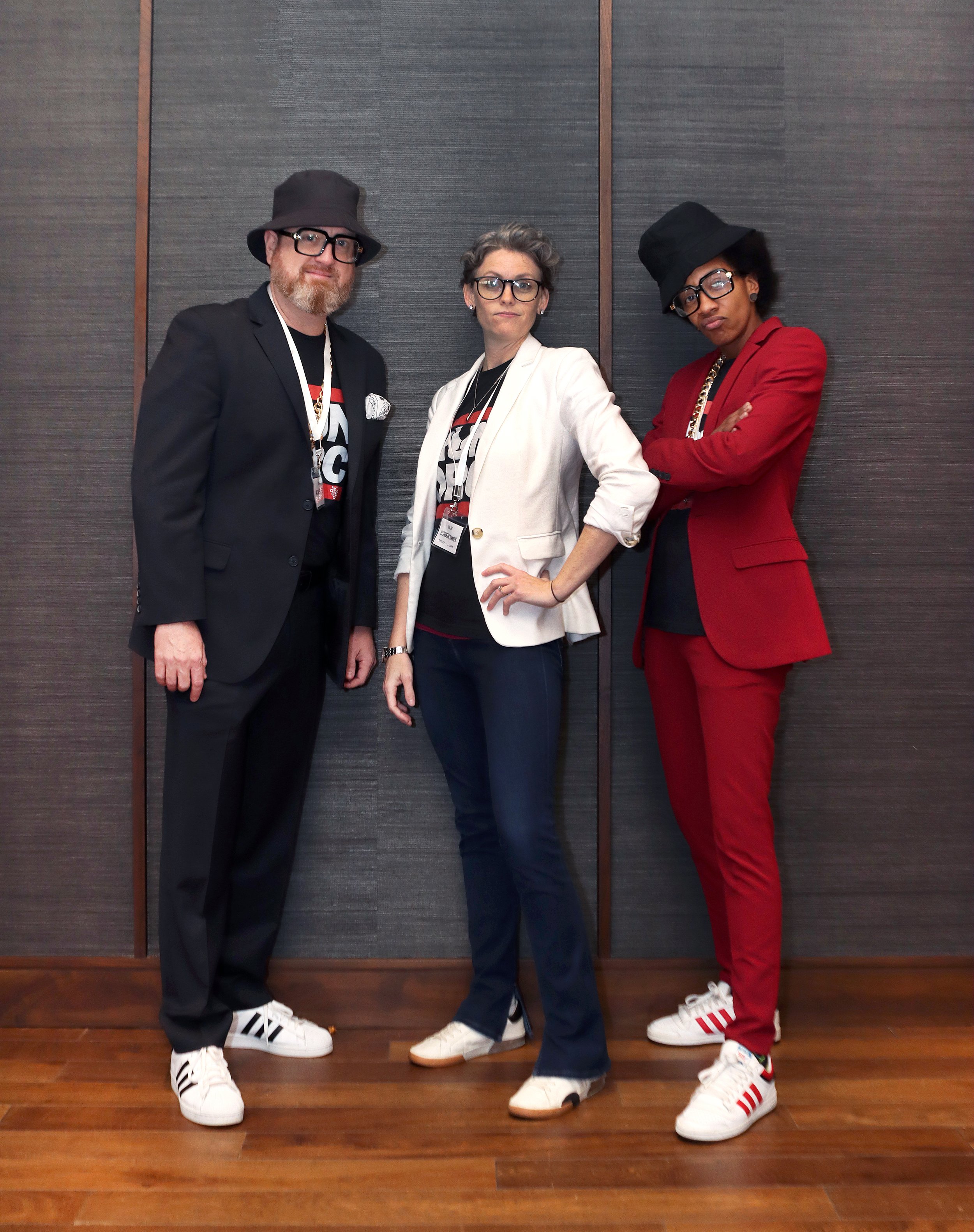 Three people standing against a wood-paneled wall. The person on the left is a man with a beard, glasses, and wearing a black blazer, a hat, and white sneakers. The person in the middle is a woman with short gray hair, glasses, and dressed in a white