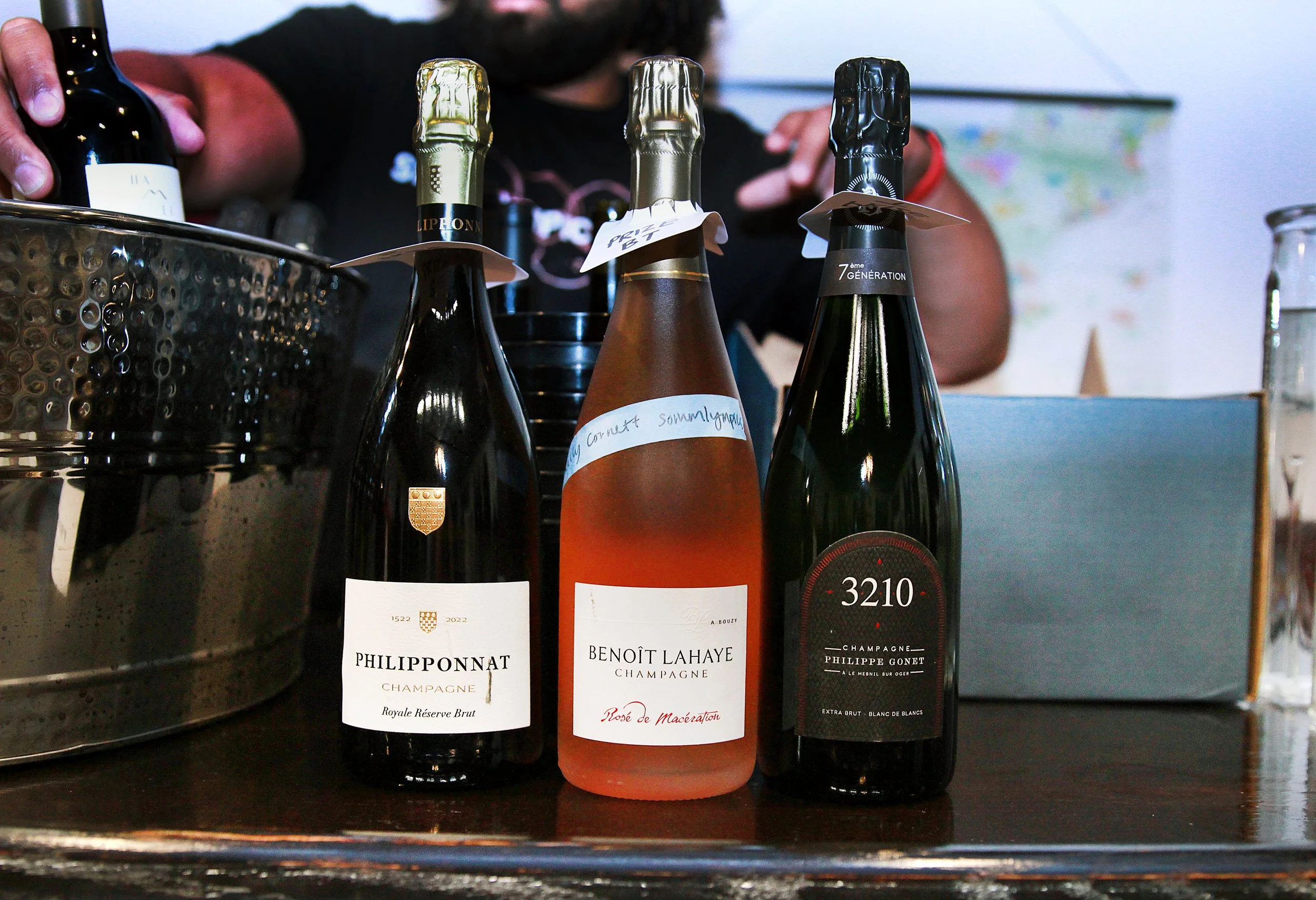 Three bottles of champagne on a table, with a person in the background. The bottles have different labels and are surrounded by other items.