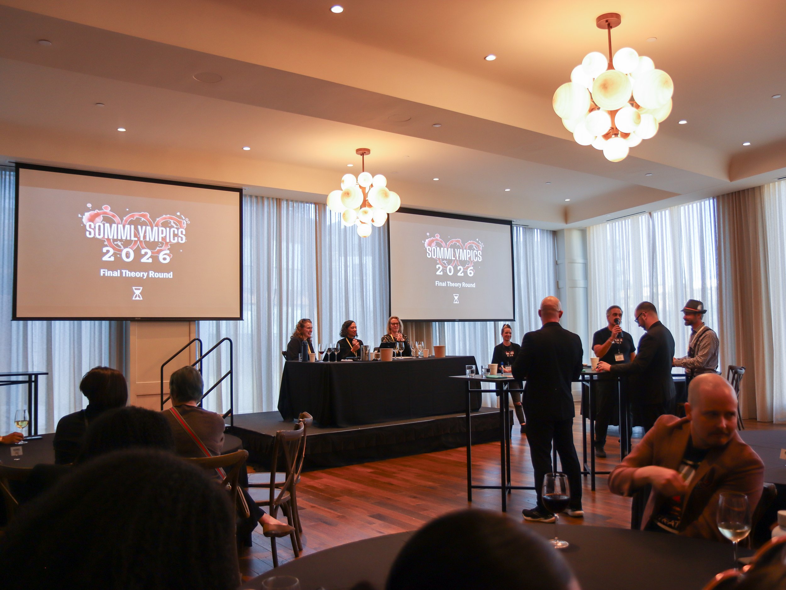 People attending an awards event called SOMMLYMPICS 2026 Final Theory Round, with two large screens displaying the event logo and name, in a well-lit conference room with chandeliers and vertical curtains.
