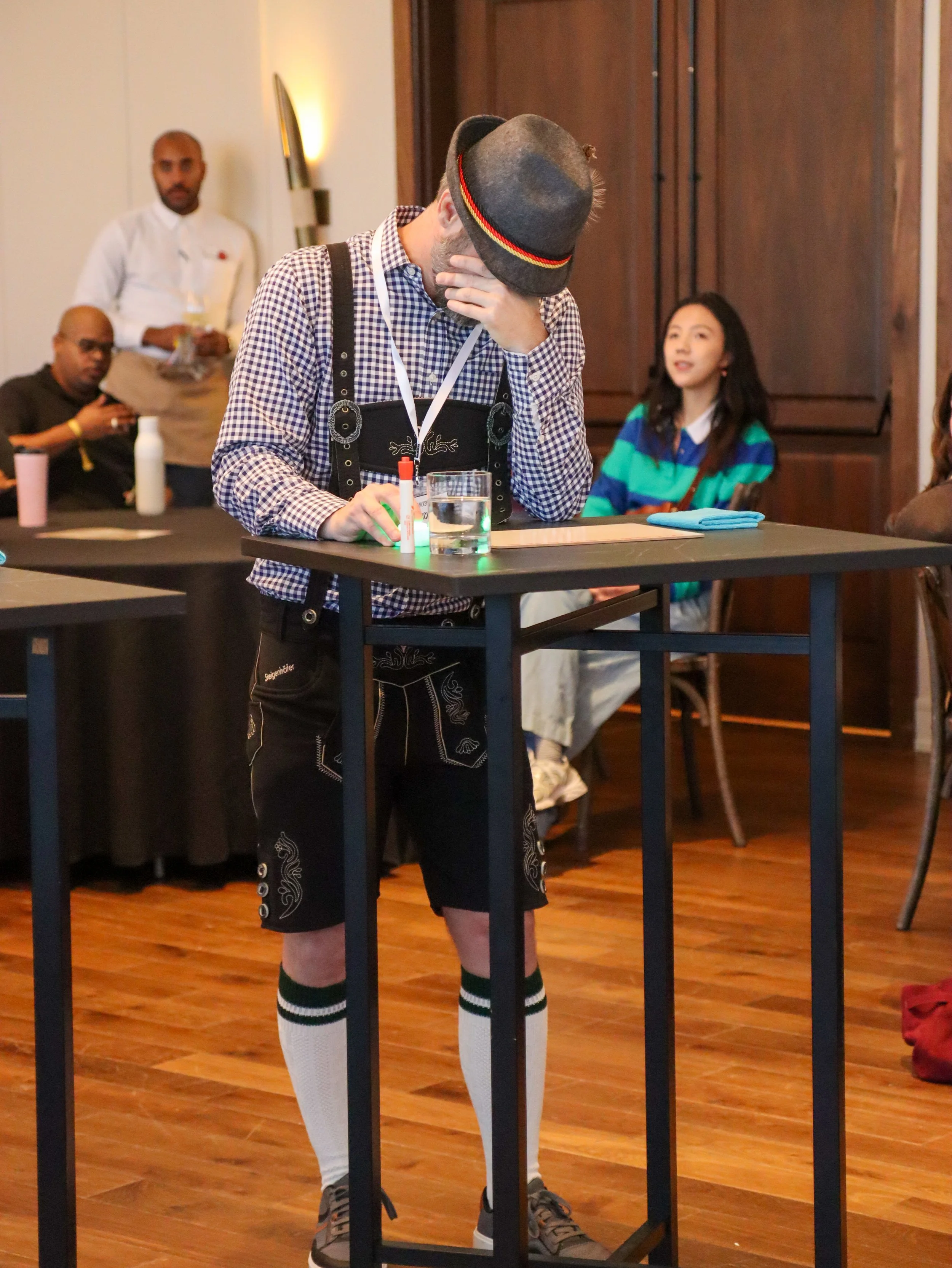 A man dressed in traditional German attire with a checkered shirt, lederhosen, and knee-high socks stands behind a table, covering his face with one hand and wearing a hat with a red, yellow, and black band. The table holds a glass of water, a small 