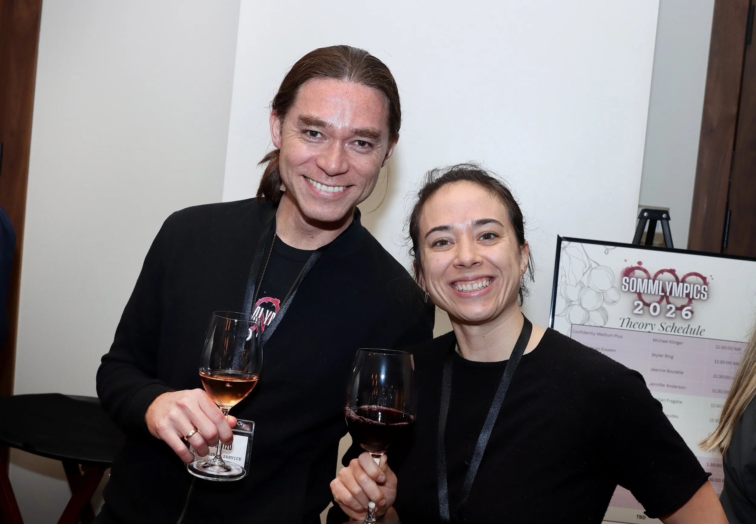 Two women at a social event, holding glasses of red wine and smiling at the camera. One woman has dark hair tied back and the other has long dark hair. There is a projector screen with a schedule and a sign that says "Sommlympics 2026" in the backgro