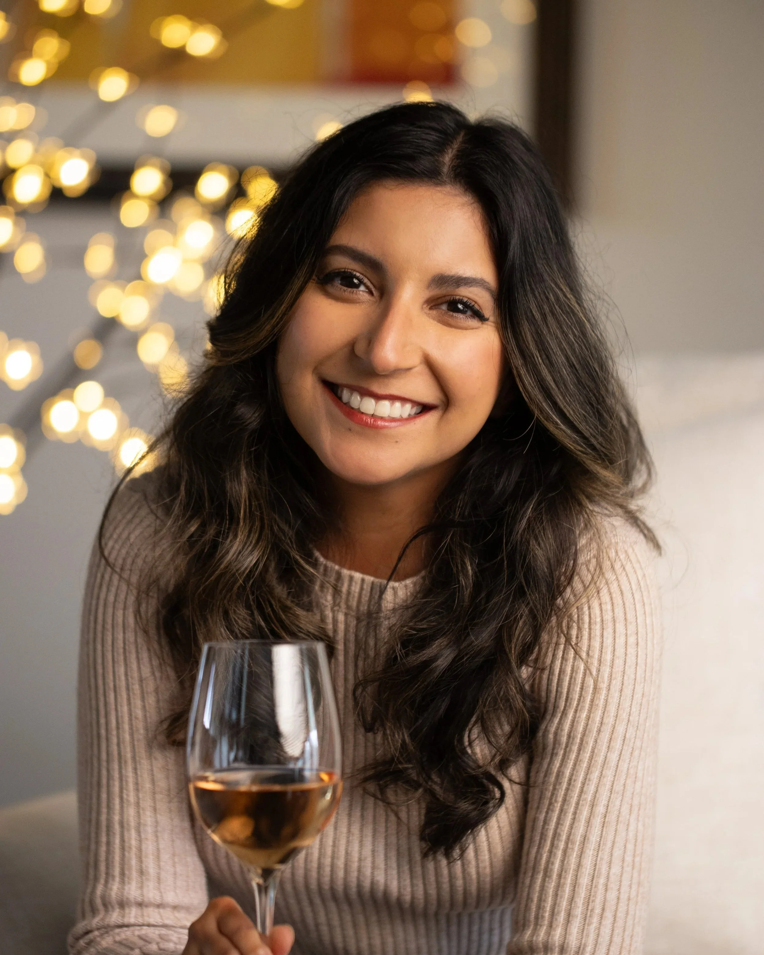 A woman with long wavy dark hair and a beige sweater smiling and holding a glass of rosé wine, with blurred warm lights in the background.