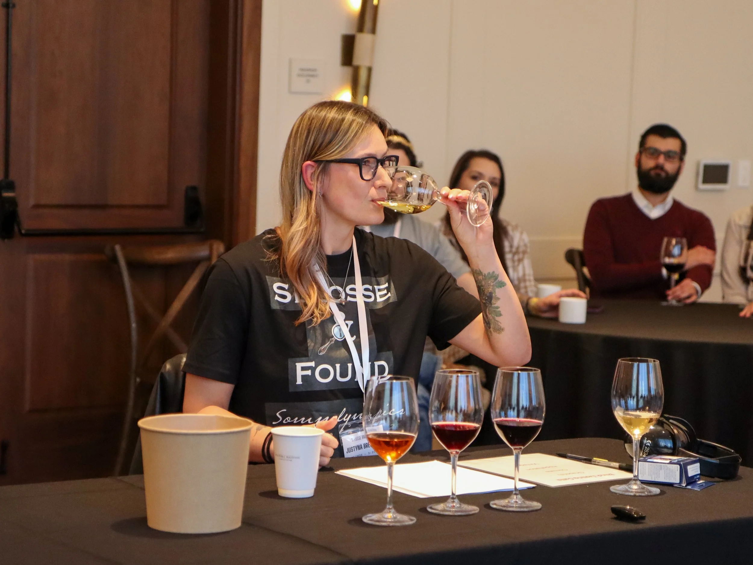 A woman with glasses is tasting wine at a wine tasting event, surrounded by glasses of various wines on the table.