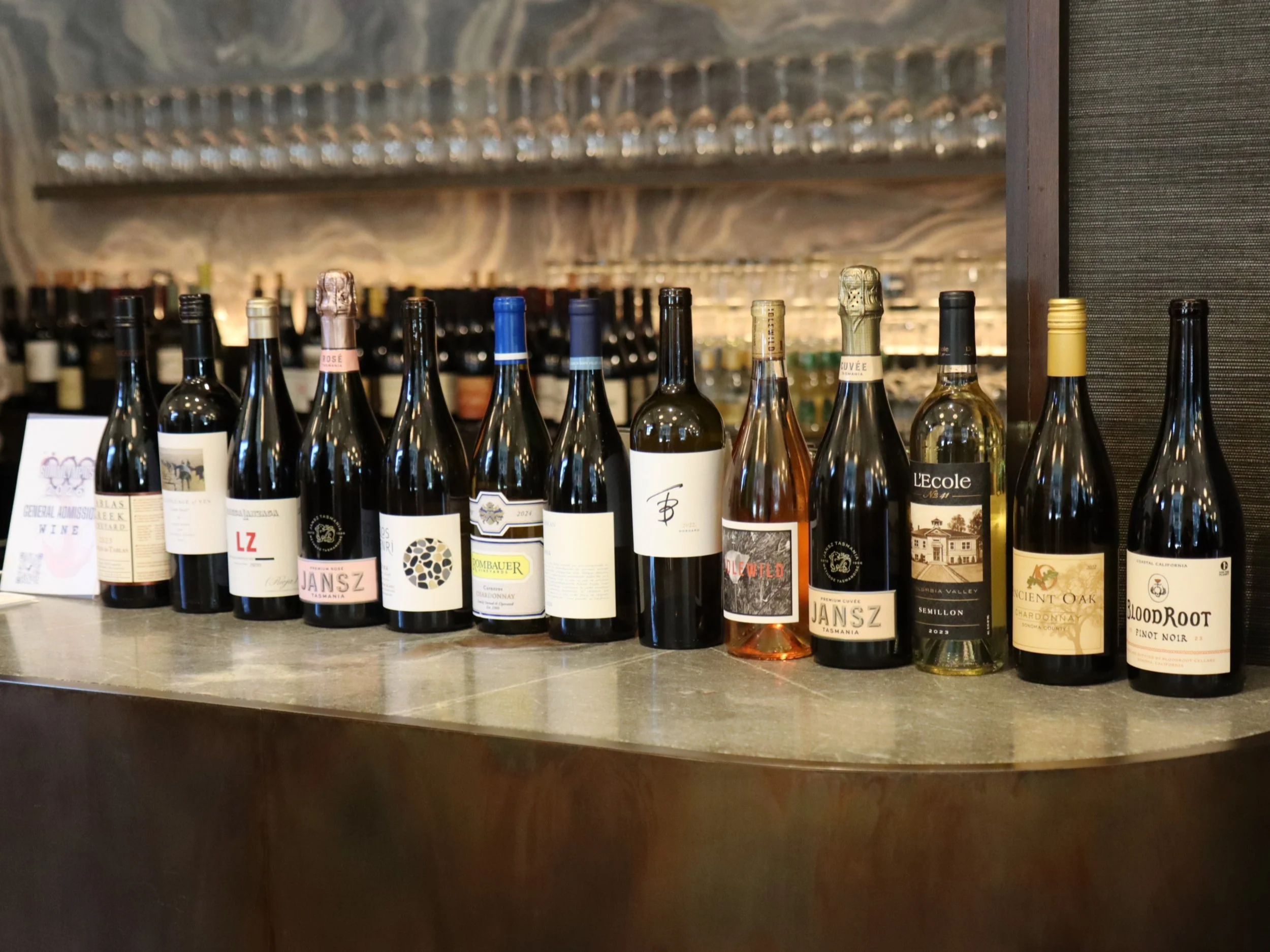 A row of wine bottles on a countertop with various labels and colors, with glassware on a wine rack above and a marble wall in the background.