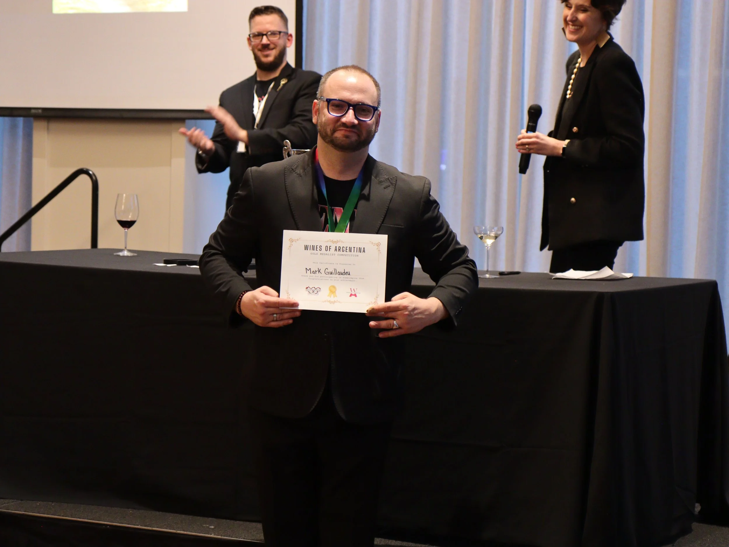 A man with glasses holding a certificate at a conference or award ceremony, with two smiling people in the background, one clapping and one holding a microphone.