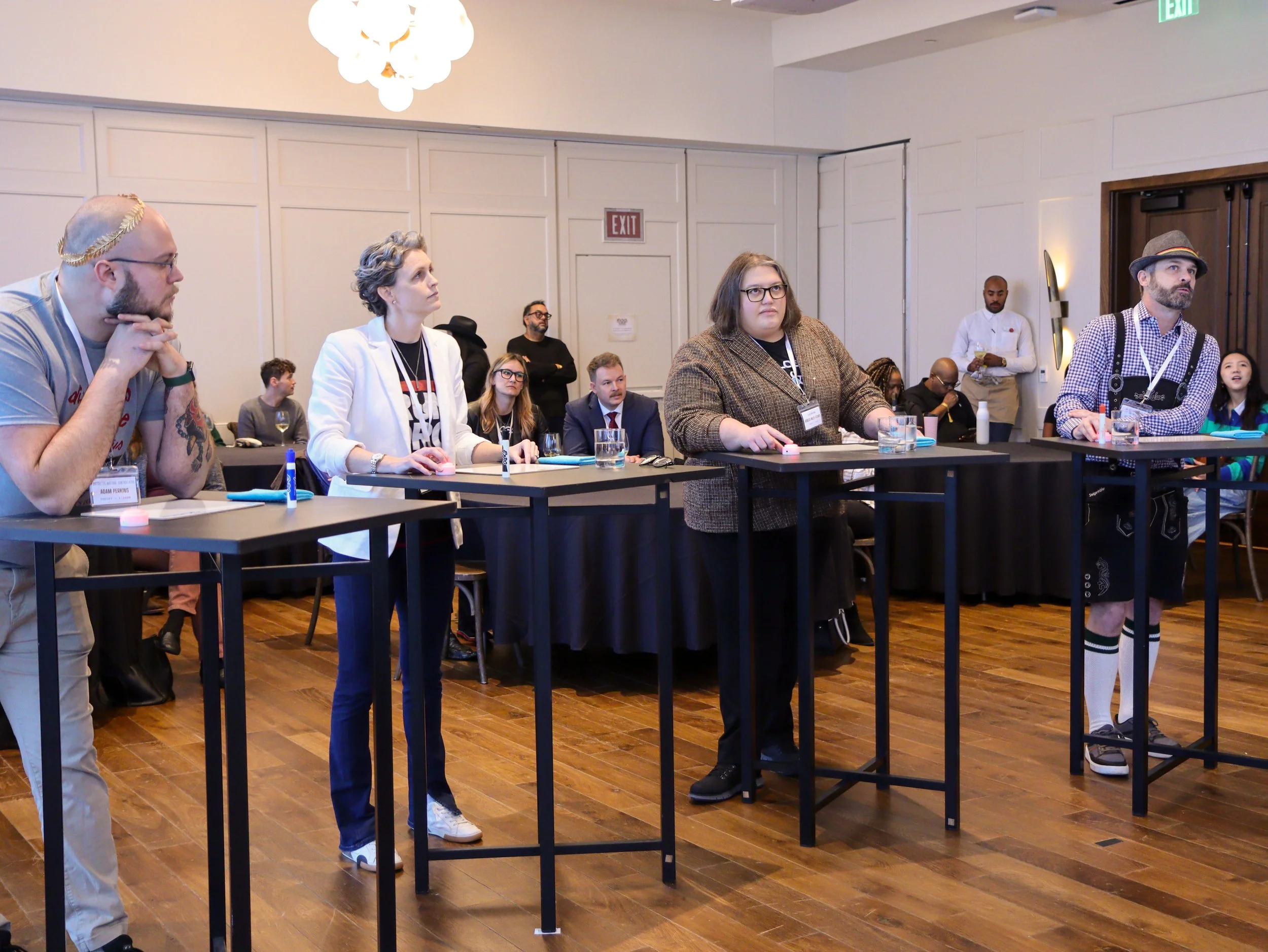 People sitting and standing around tables in a conference room during a meeting or workshop. Several individuals are attentively listening or engaging, with drinks and writing materials on the tables. An exit sign is visible on the wall.