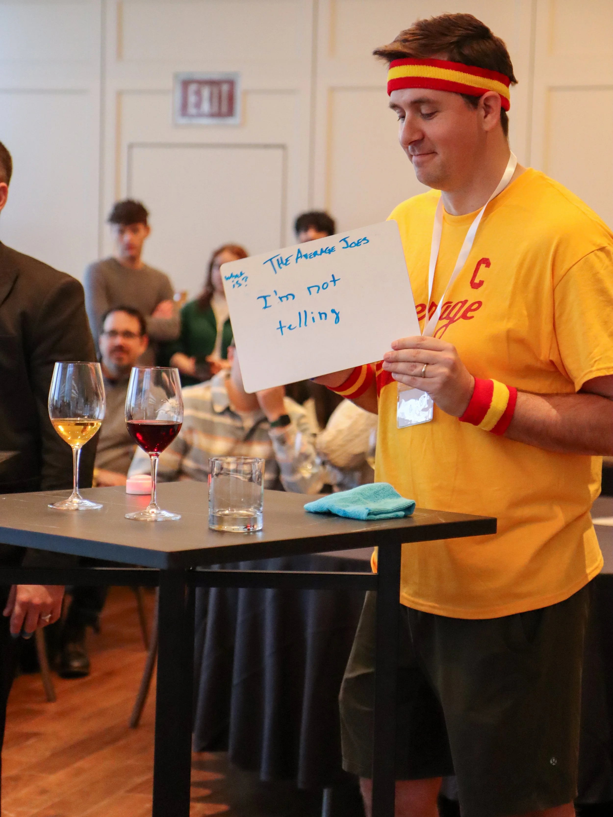 A man wearing a yellow shirt, red wristbands, and a red and yellow headband holding a whiteboard with writing, standing at a table with glasses and a blue cloth, in a room with seated and standing people in the background.