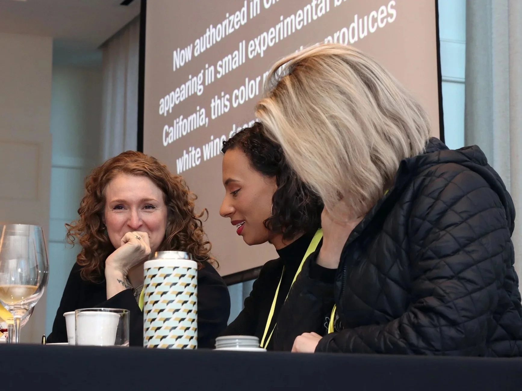 Three women sitting at a table with drinks, engaged in conversation, with a large presentation screen displaying text in the background.