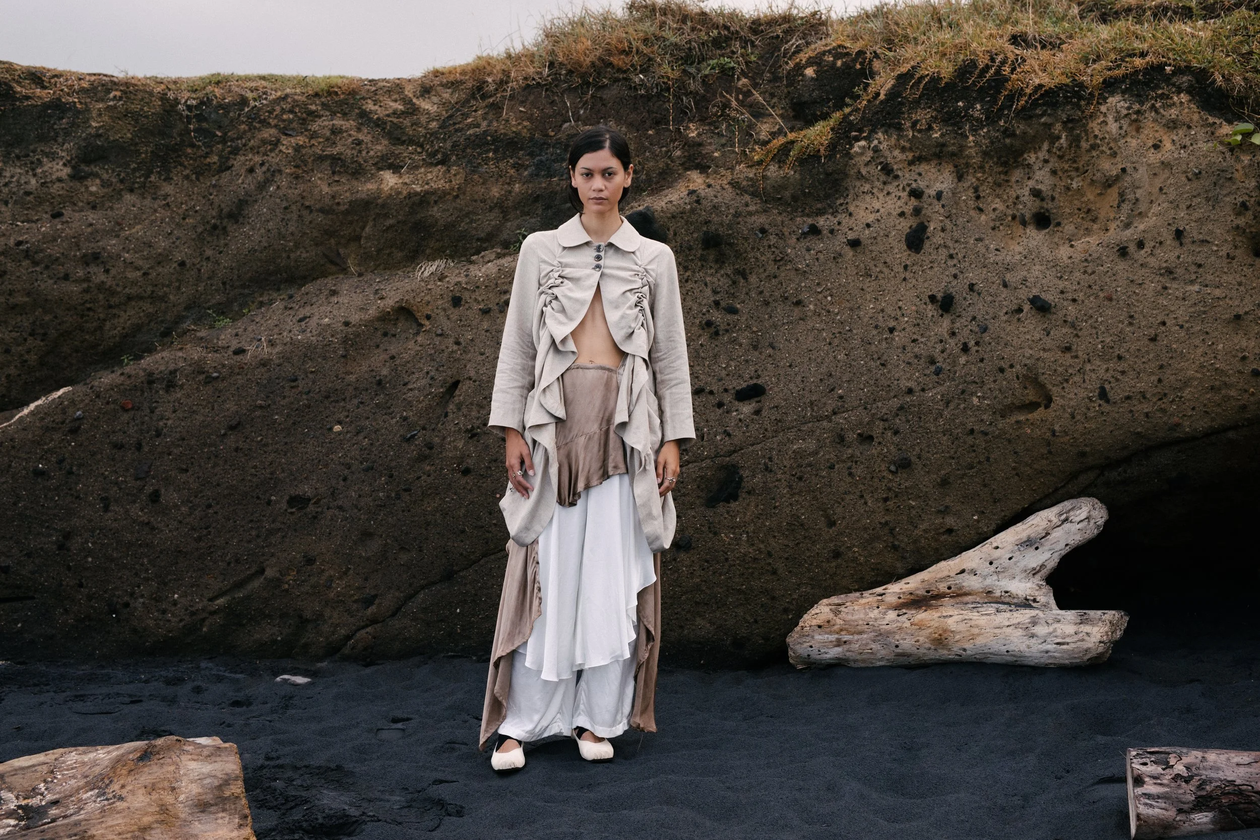 Young woman standing on dark sand near large rocks and a rocky outcrop, wearing a layered beige and white fashion outfit.