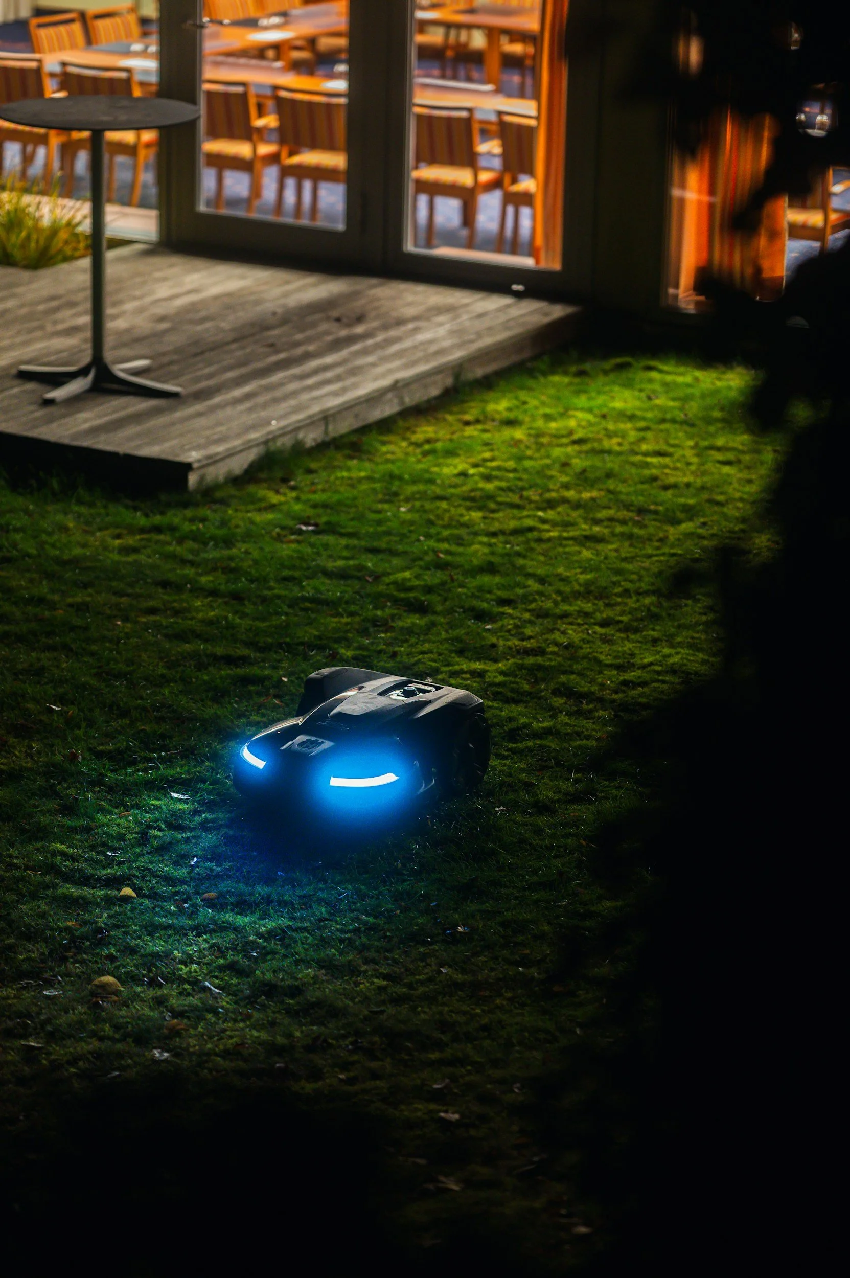 A remote-controlled robot with blue lights on a grassy lawn near a house with a deck and patio furniture.