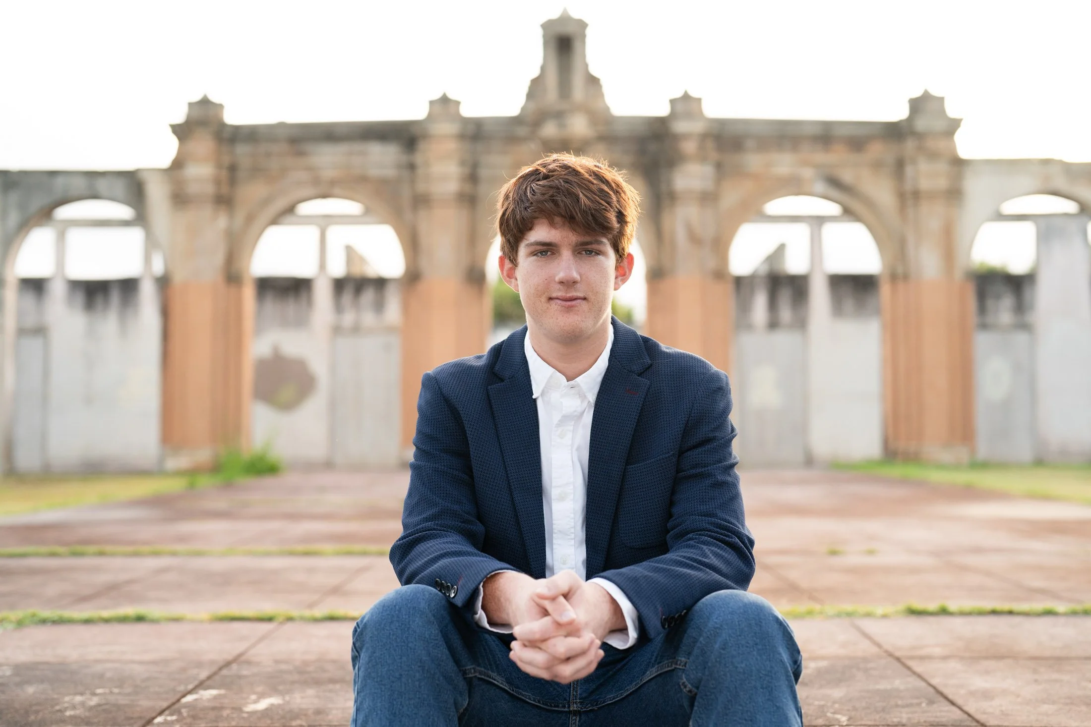 A young man with short brown hair wearing a navy blazer and white shirt, sitting on a paved area with a historic building with arches in the background for his branding photoshoot maui.