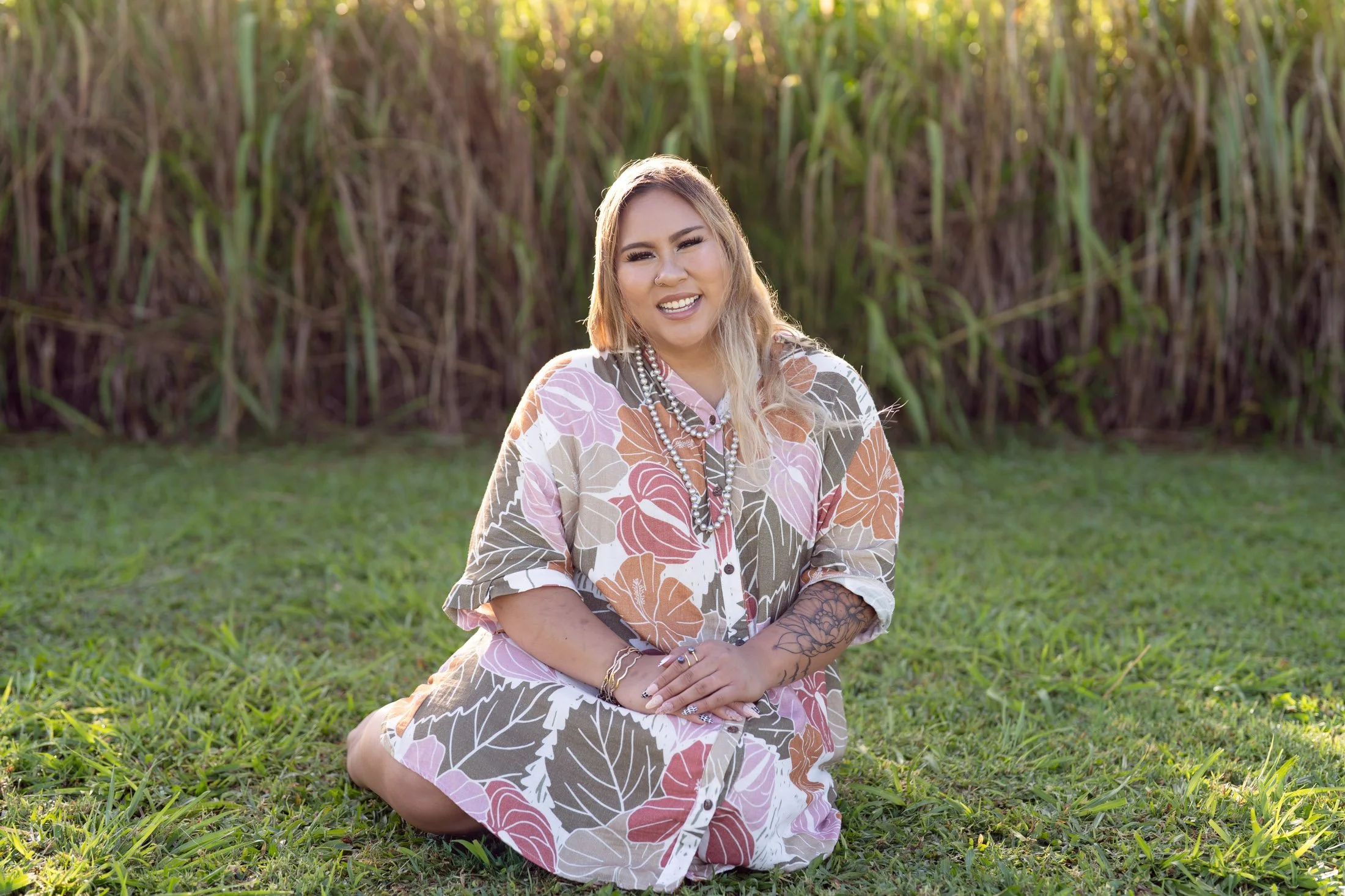 A smiling woman sitting on the grass in front of tall green plants, wearing a floral dress and jewelry for her creative business portraits.