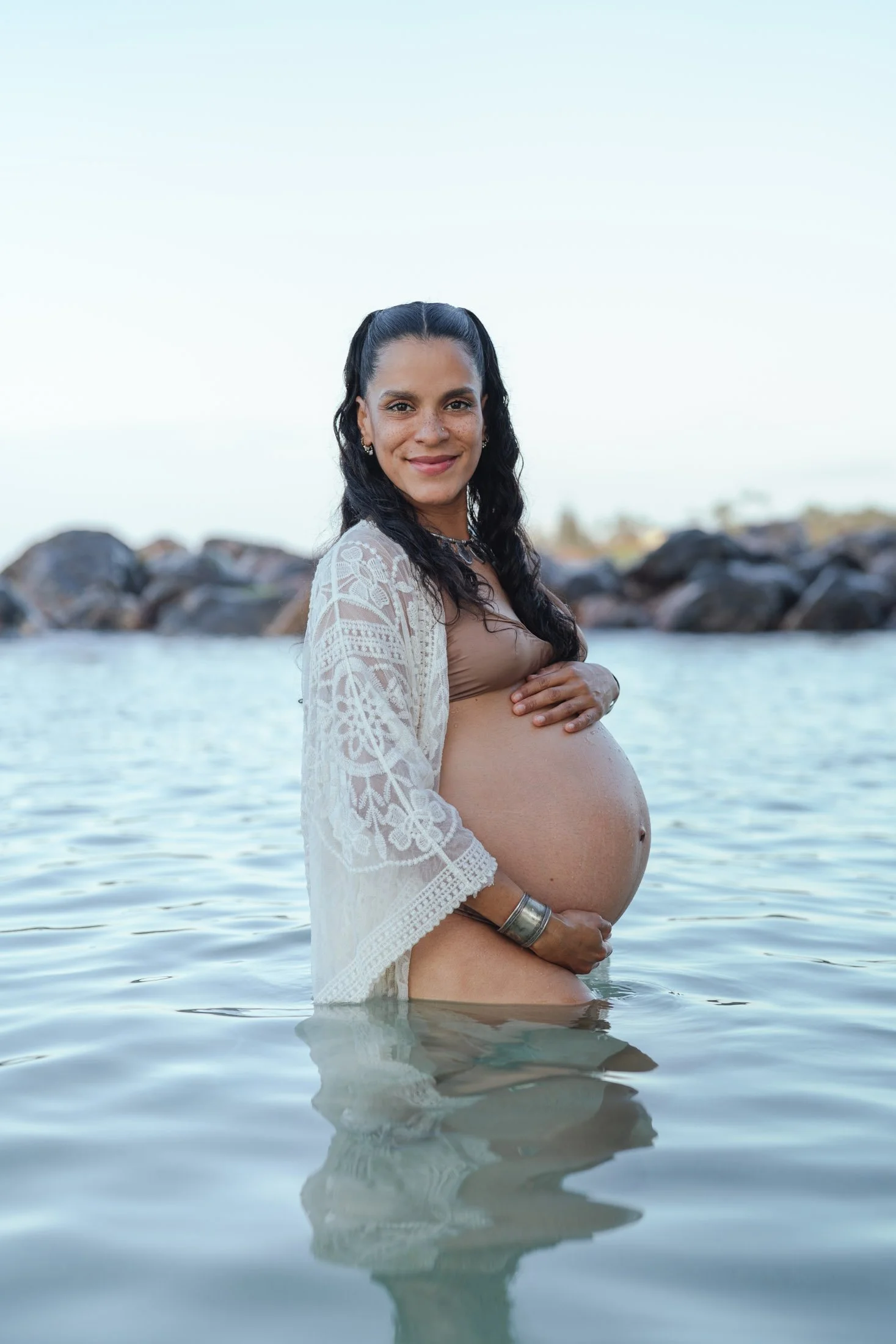 A pregnant woman standing in water at the beach, smiling and holding her belly, wearing a beige top and a white lace cover-up during her intimate portrait photography session. 