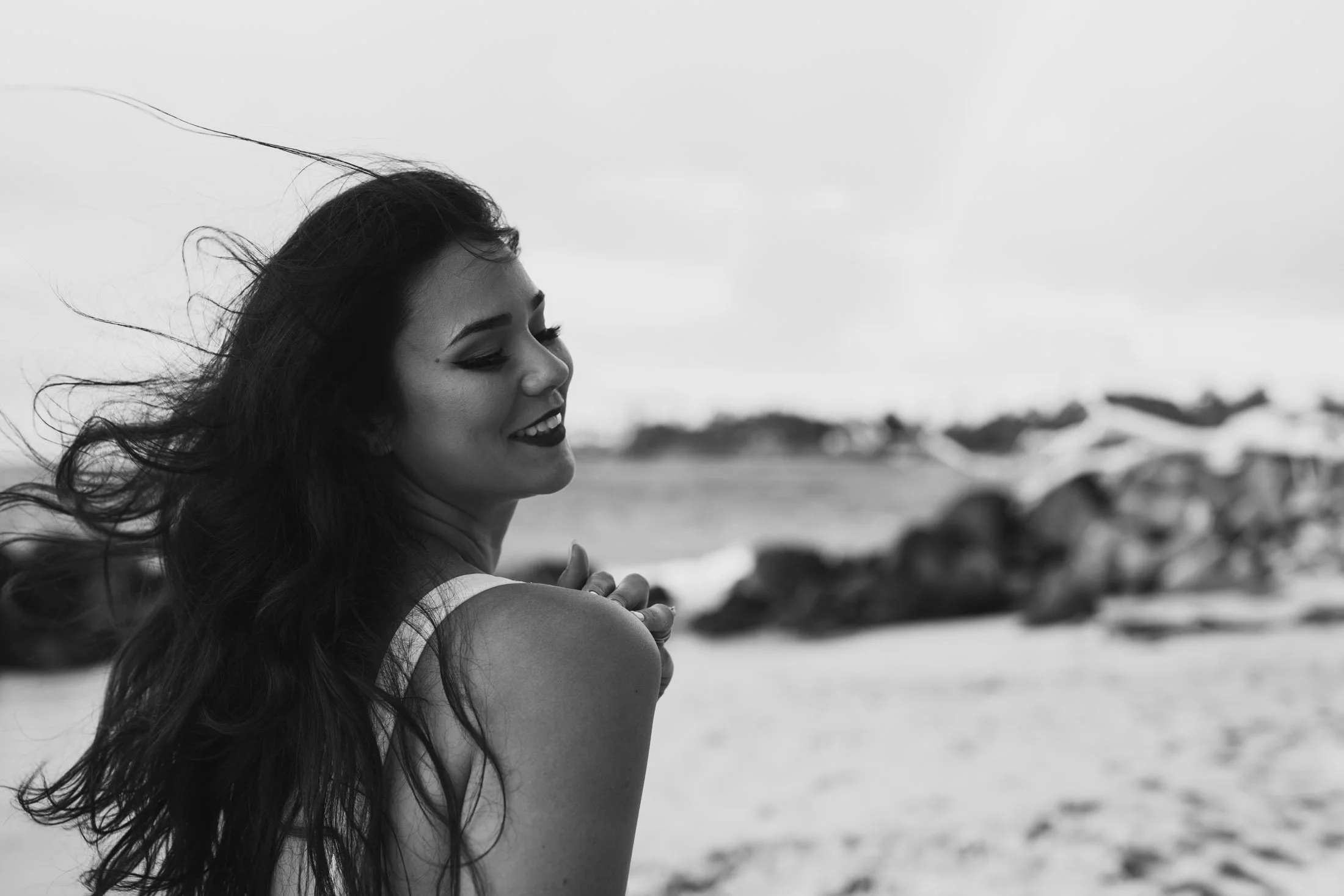 A smiling woman with long hair on a beach, with waves and rocks in the background, black and white photo for her natural light portraits maui.