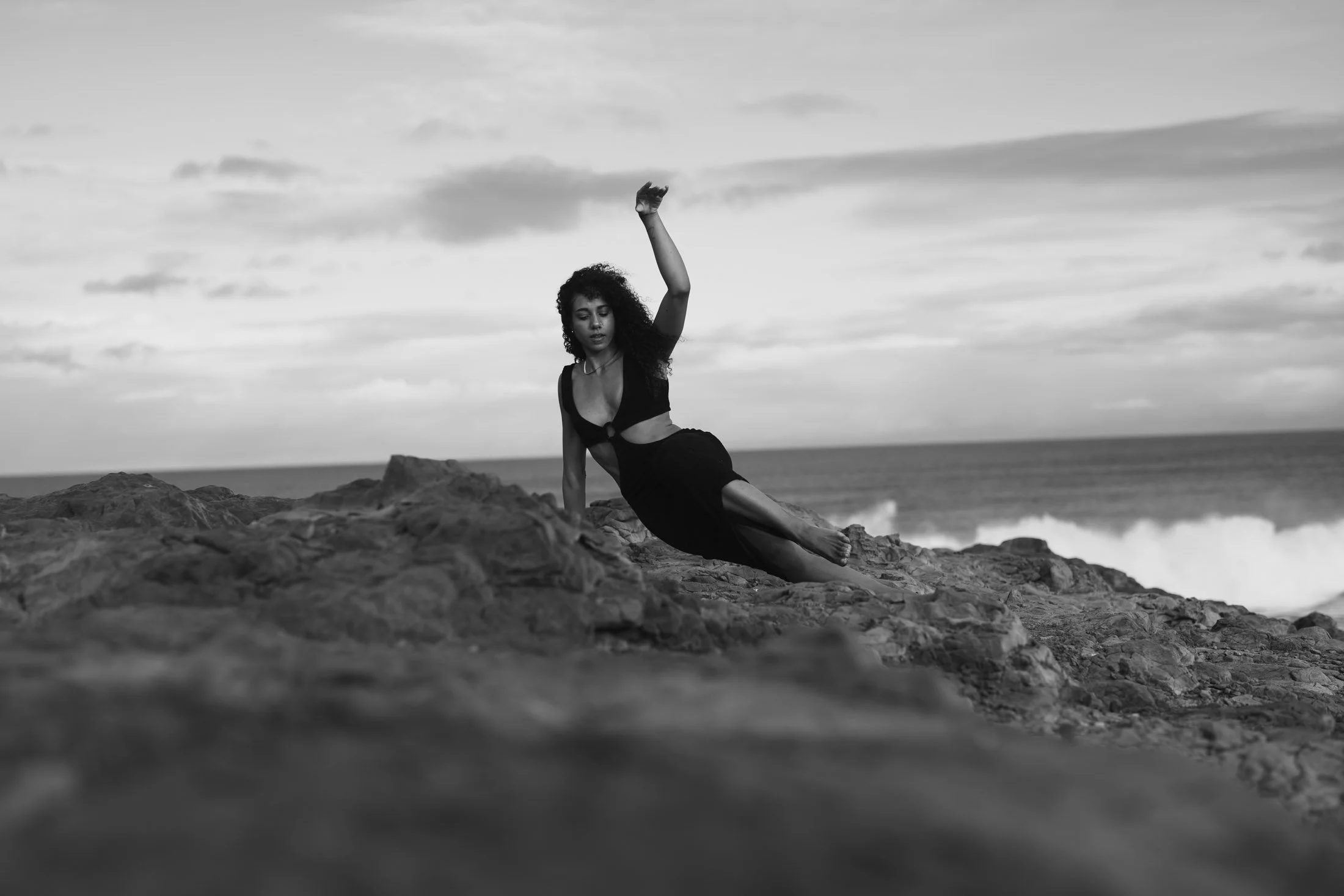 A woman with curly hair wearing a black cut-out dress is posing on a rocky beach with ocean waves and cloudy sky in the background for her outdoor portrait photography.