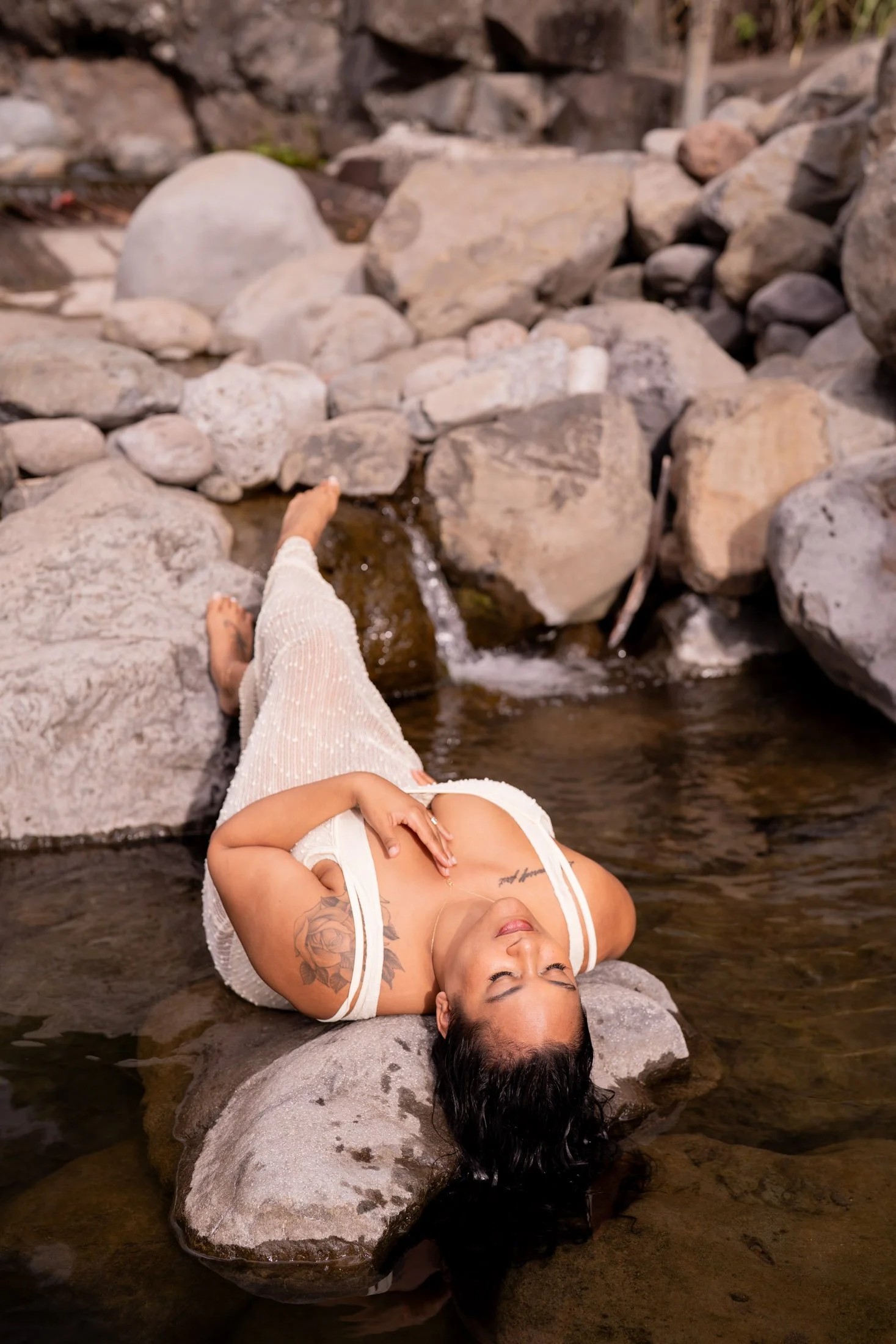 A woman with tattoos relaxing in a shallow stream surrounded by rocks for her outdoor boudoir photography session.