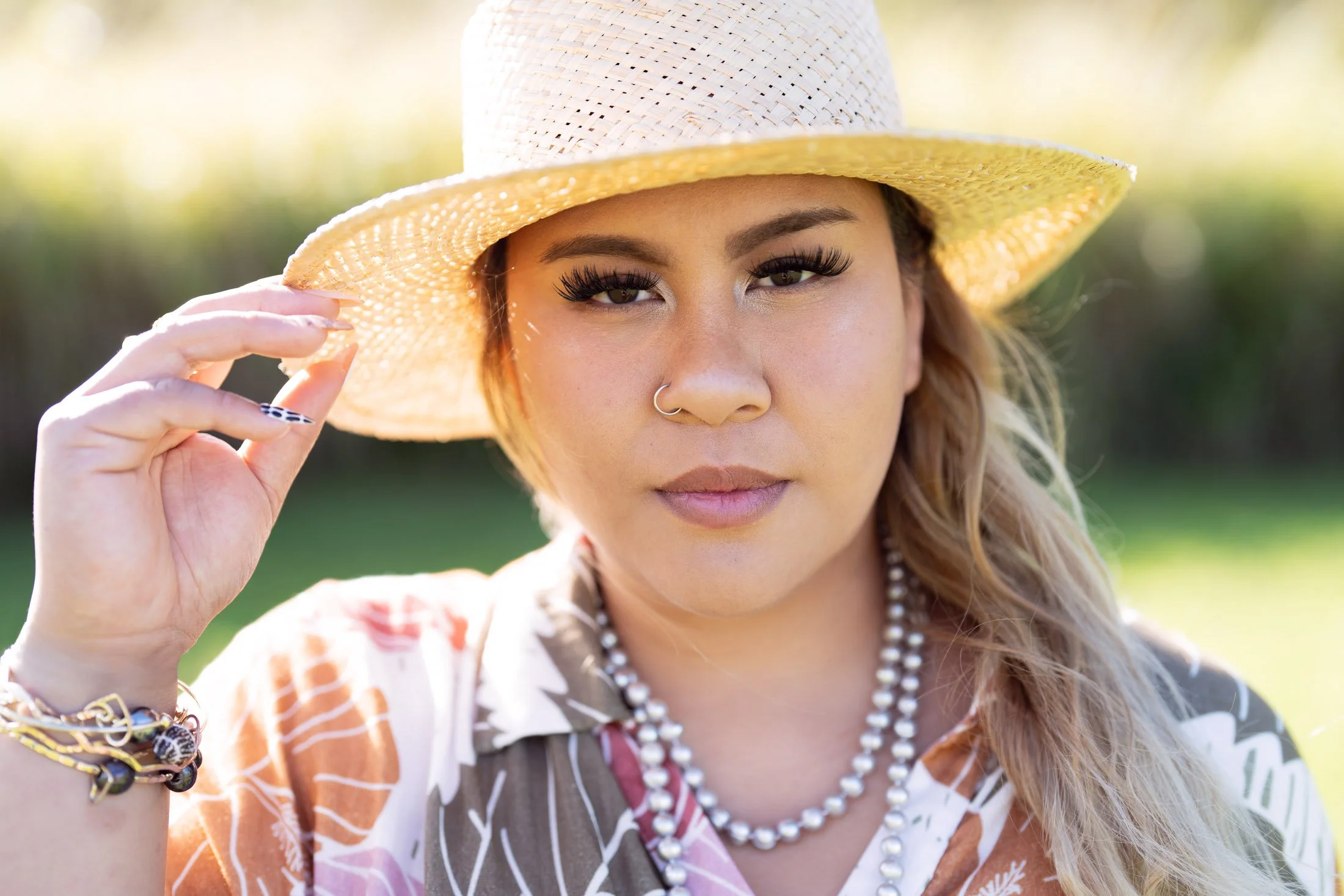 A woman wearing a straw hat, pearl necklace, and colorful patterned shirt, looking at the camera outdoors in bright sunlight during her entrepreneur photoshoot.