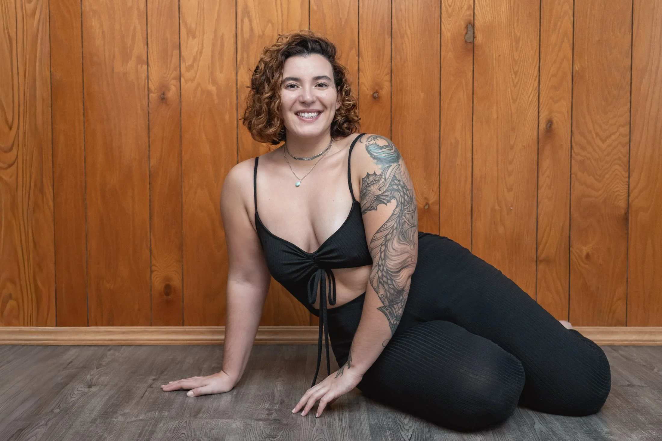 A woman with curly hair, wearing a black crop top and leggings, smiling and sitting on the floor against a wooden wall, with tattoos on her arm for her entrepreneur photoshoot.
