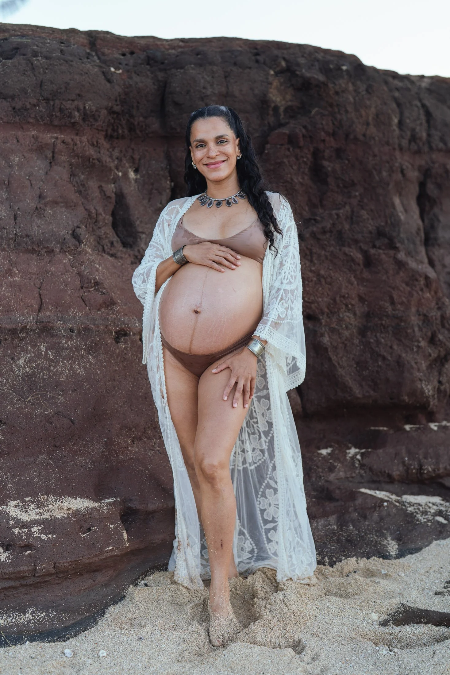 Pregnant woman standing on sandy beach with large rocks behind her, wearing a brown bikini, white lace cover-up, and jewelry, smiling at the camera for her artistic photoshoot in maui.