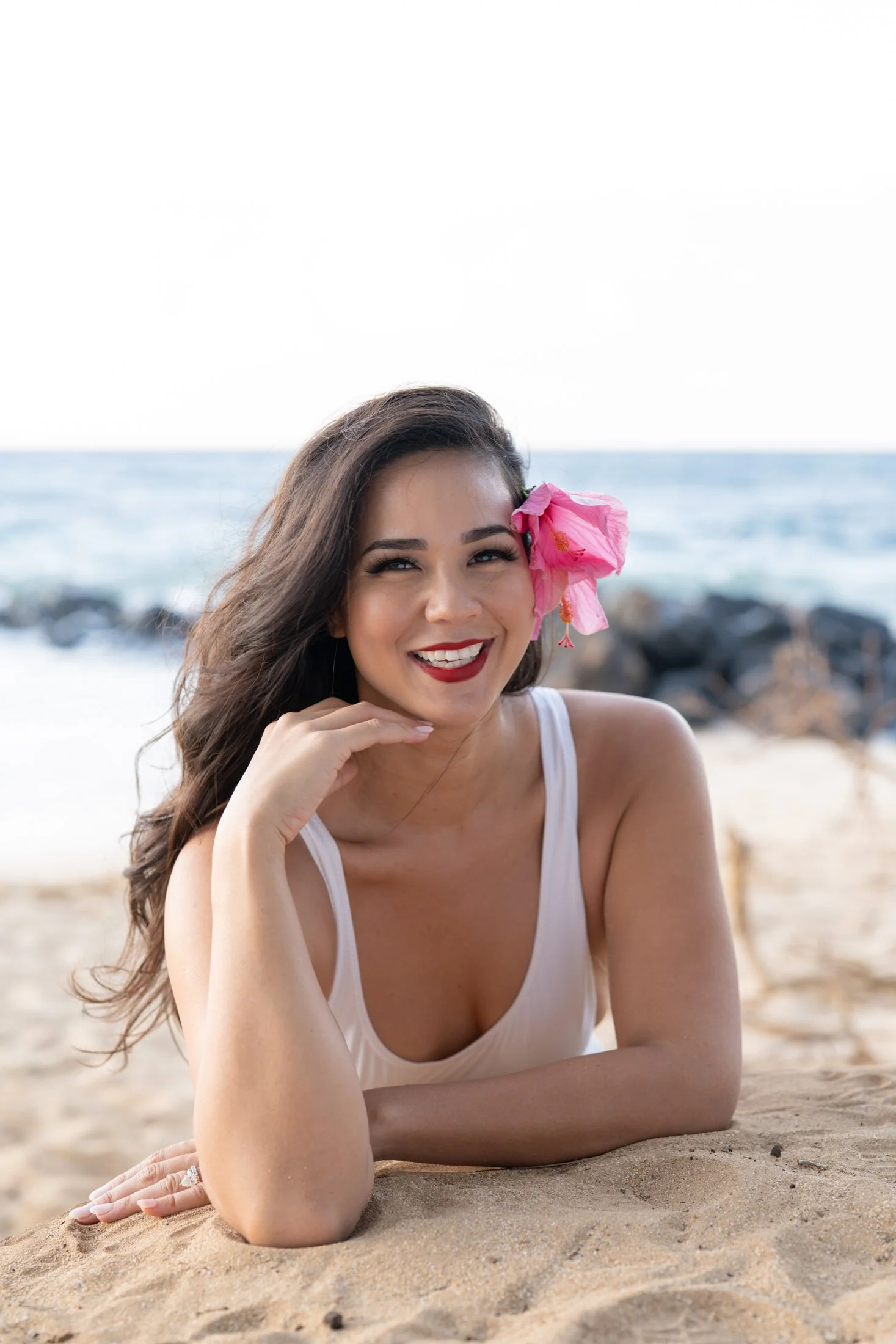 A smiling woman lying on the beach with a pink flower behind her ear during her outdoor portrait photography session.